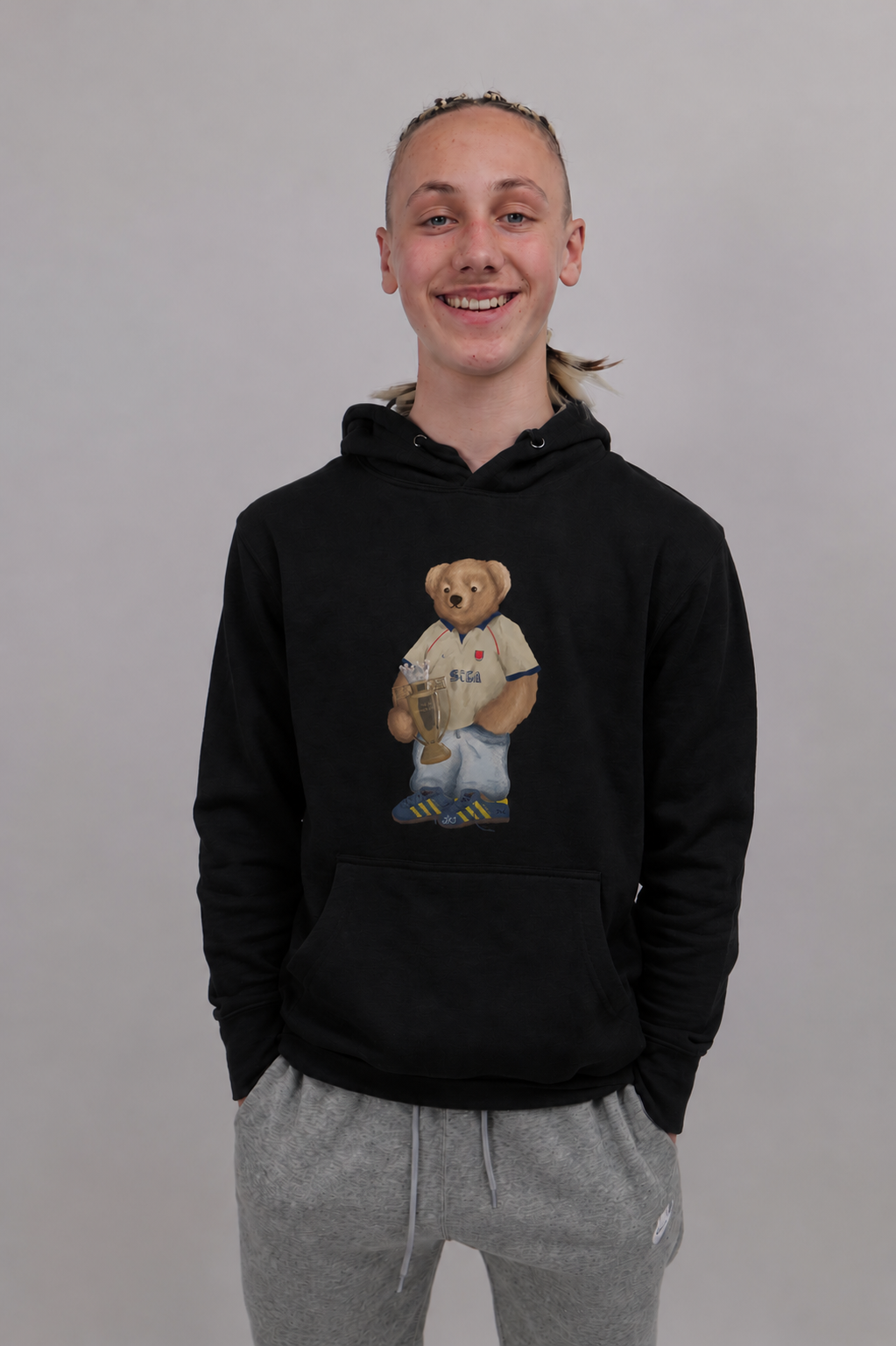 A smiling young man wearing a black pullover hoodie with a teddy bear graphic holding a trophy on the chest, paired with gray sweatpants, against a plain gray background.