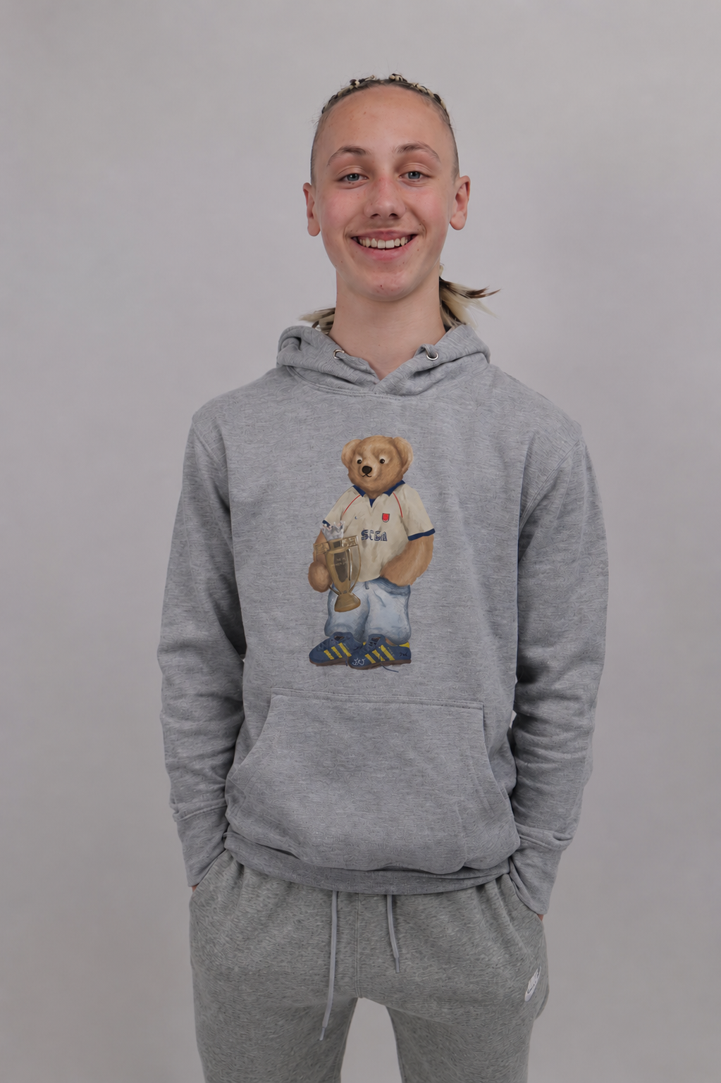 A smiling young man wearing a heather gray pullover hoodie with a teddy bear graphic on the chest, paired with matching gray sweatpants, against a plain light gray background.