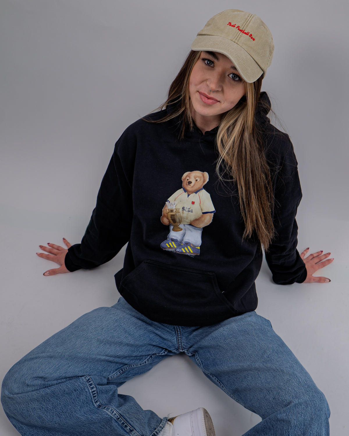 Person wearing a black sweatshirt with a bear graphic and a beige cap, sitting on a white surface.