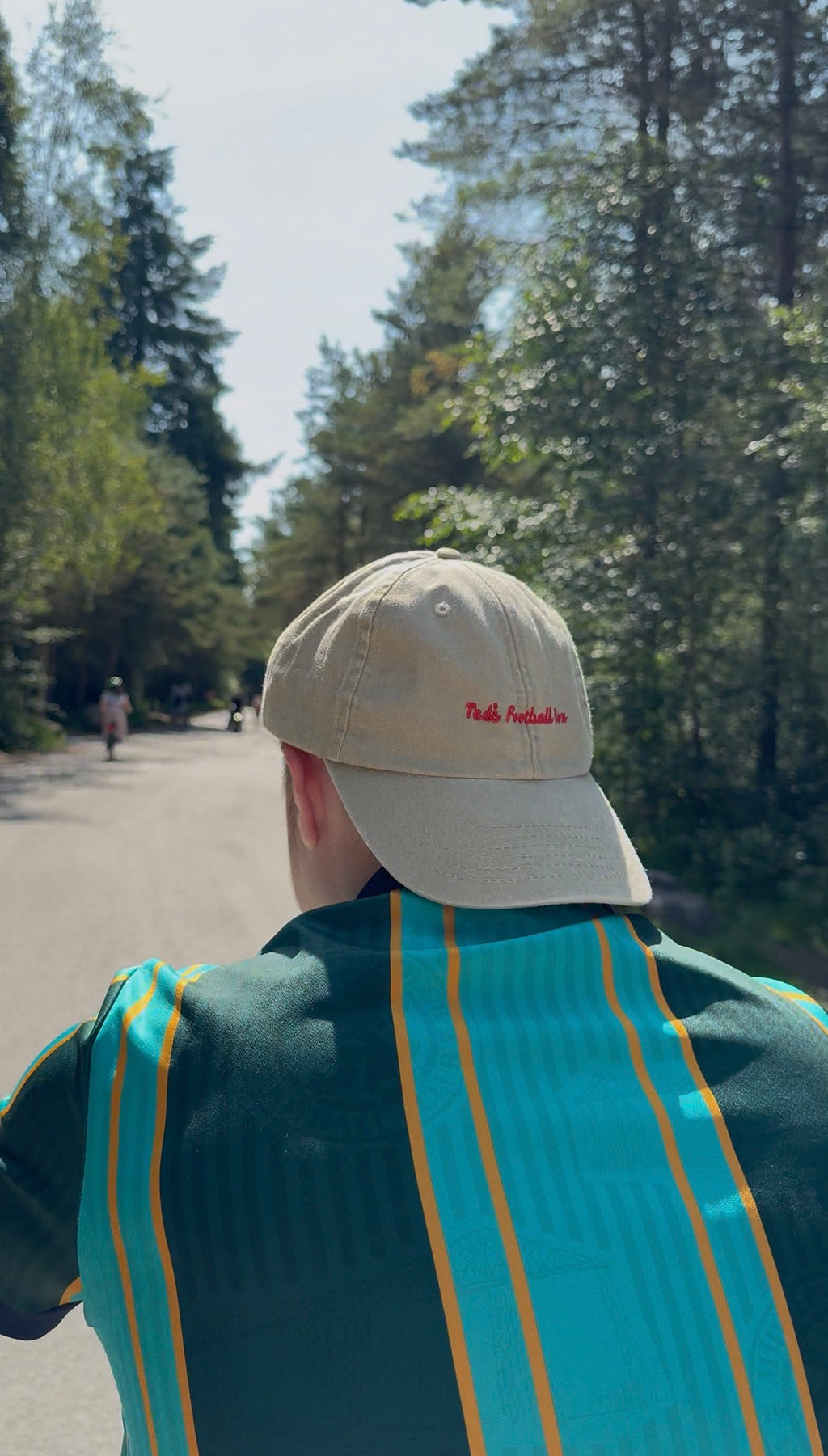 Person wearing a beige cap with text, walking on a path with trees in the background