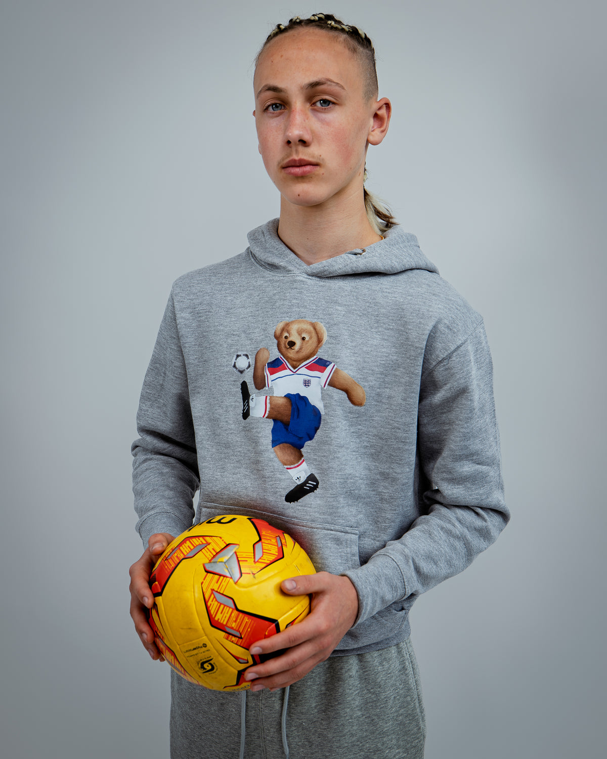 Person wearing a gray hoodie with a bear graphic holding a soccer ball against a gray background