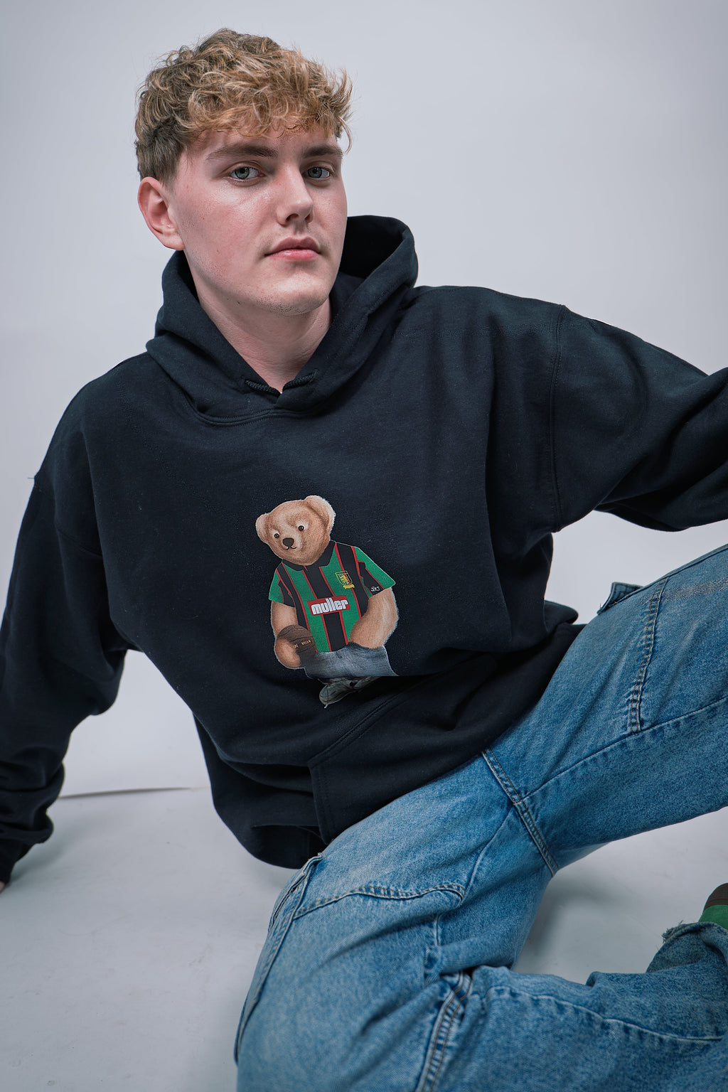 Person wearing a black hoodie with a bear graphic, sitting on a white floor against a light gray wall.