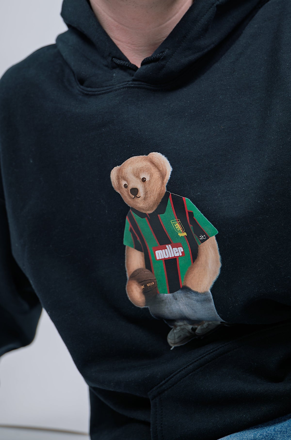 Black hoodie with a teddy bear wearing a striped shirt and holding a soccer ball on a white background