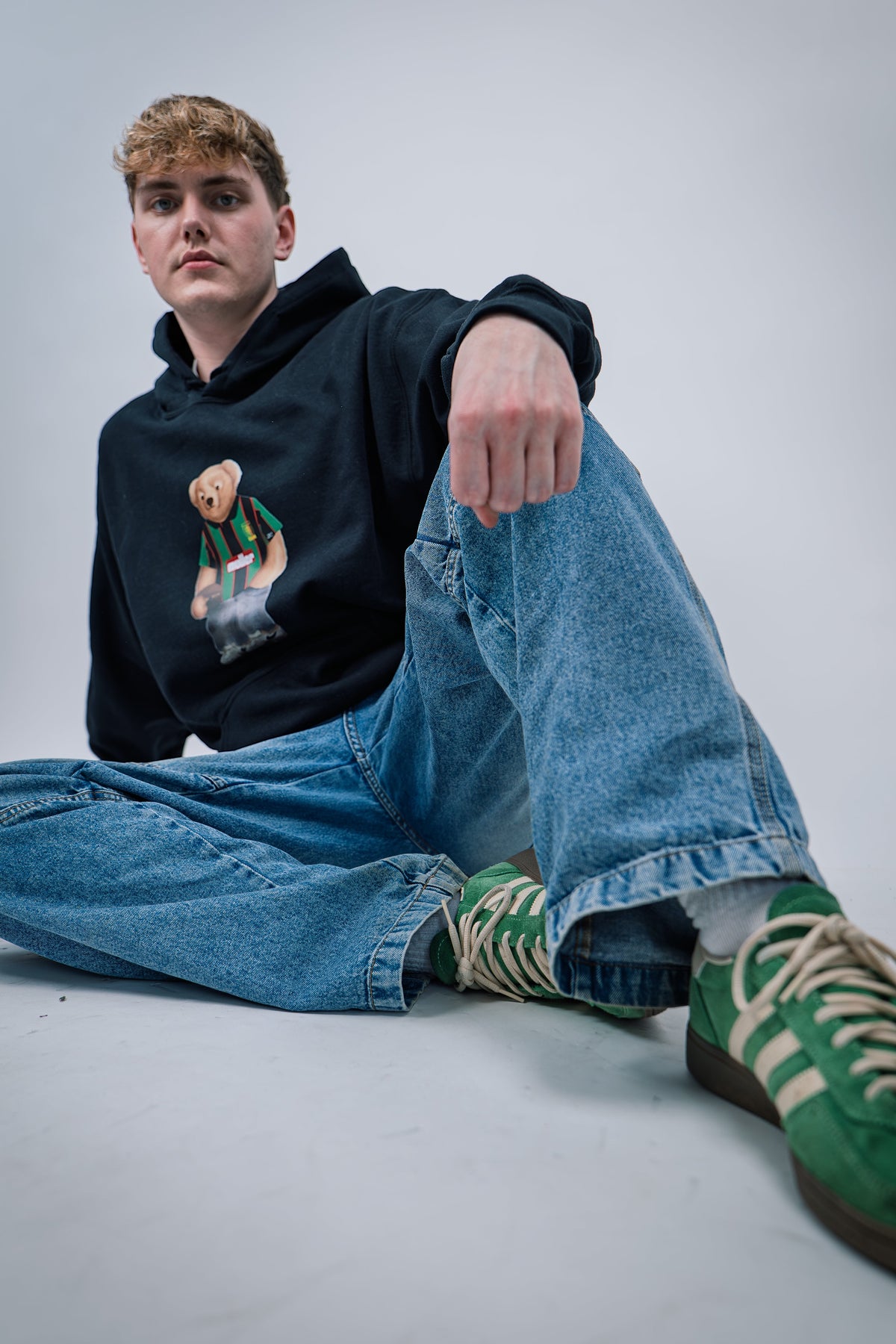 Person wearing a black hoodie with a graphic design, blue jeans, and green sneakers sitting on a white background.