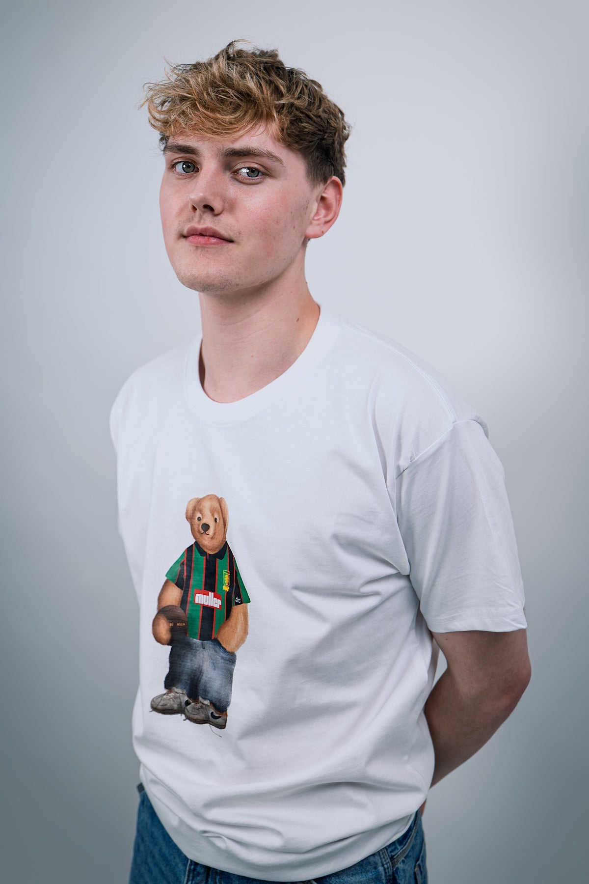 Person wearing a white t-shirt with a graphic of a Aston Viilla bear in a striped sweater on a plain background