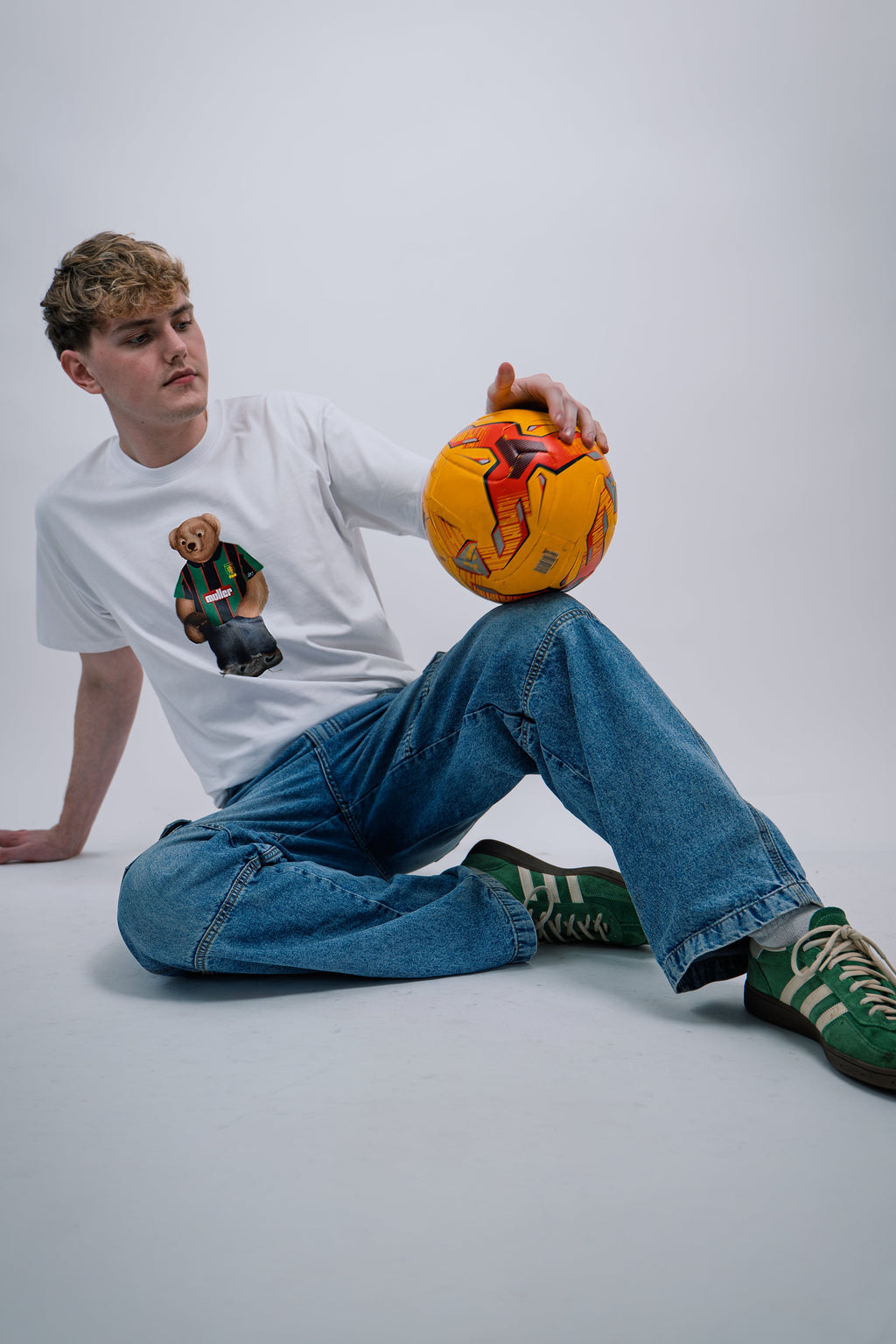 Person wearing a Aston villa white tshirt sitting on the floor holding a soccer ball against a plain background