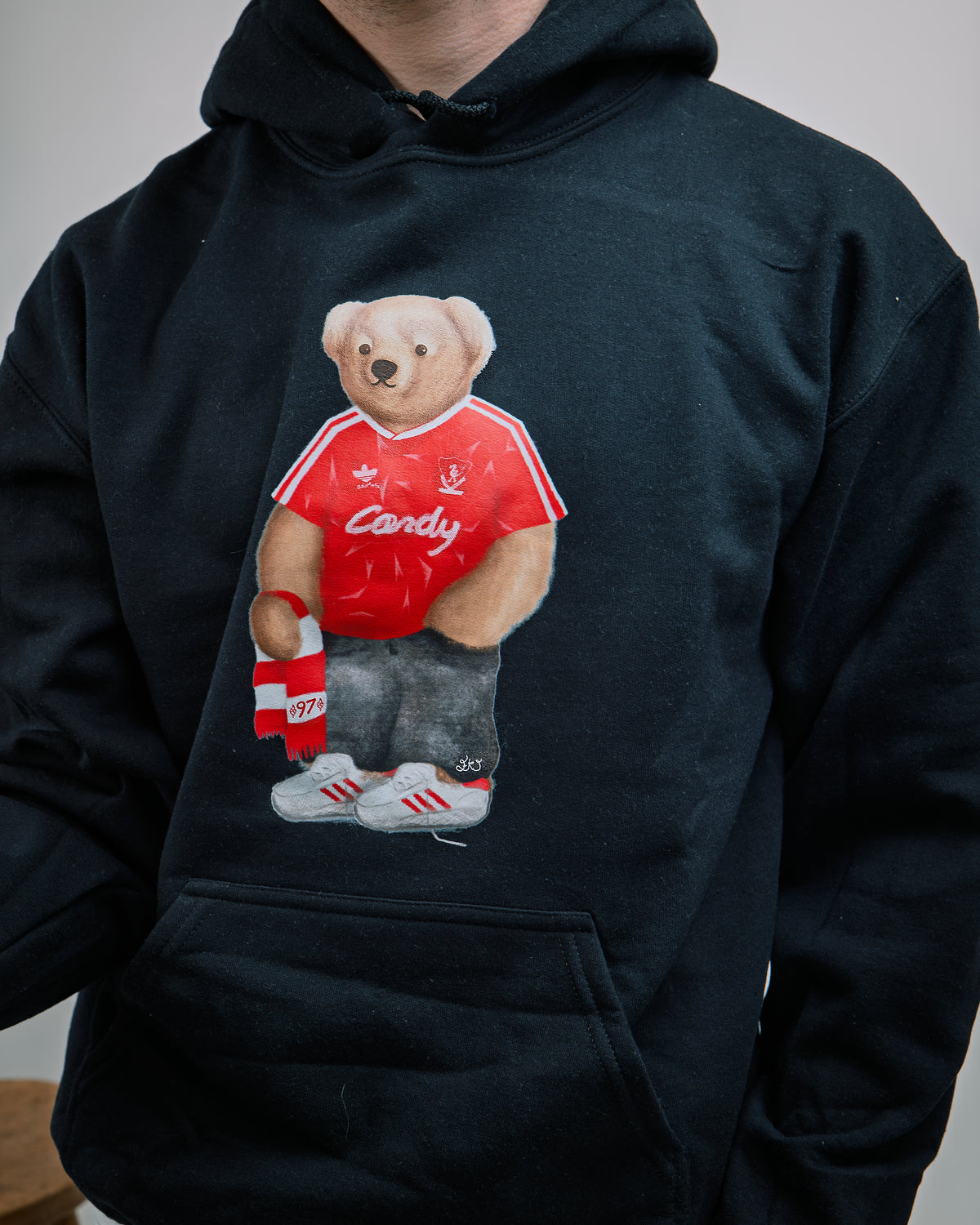 Black hoodie with a bear graphic wearing a red shirt and Adidas shoes on a plain background