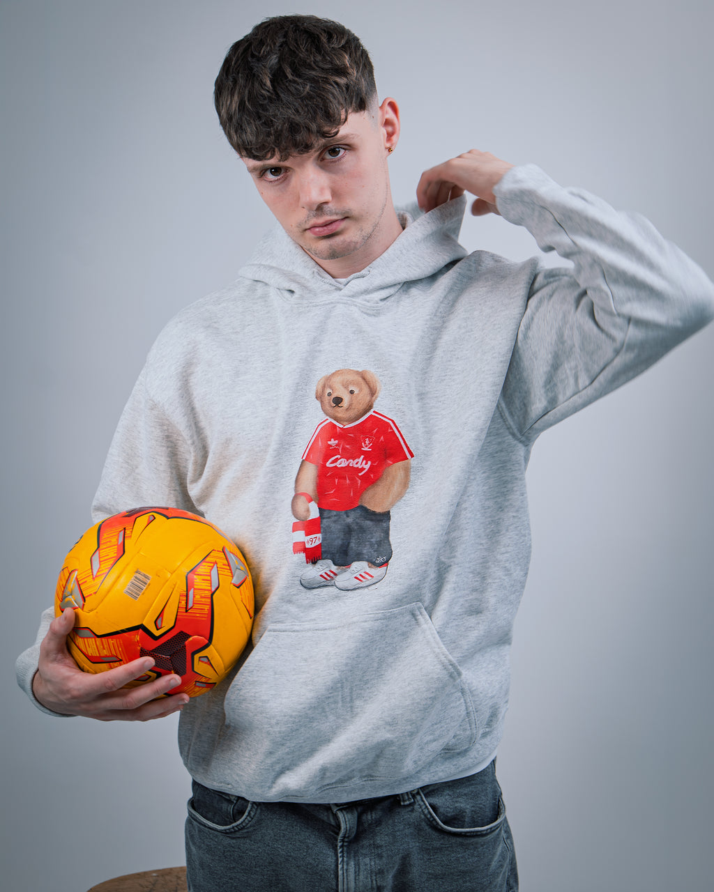 Person wearing a hoodie with a teddy bear graphic holding an orange soccer ball against a gray background