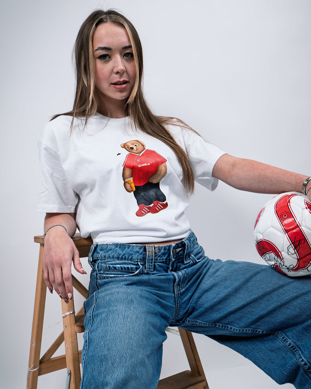 Person wearing a white sweatshirt with a graphic design, sitting on a wooden stool holding a soccer ball.