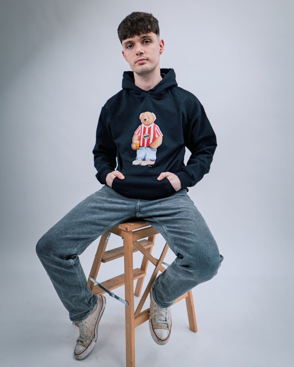 Person wearing a black hoodie with a teddy bear graphic, sitting on a wooden stool against a plain background.