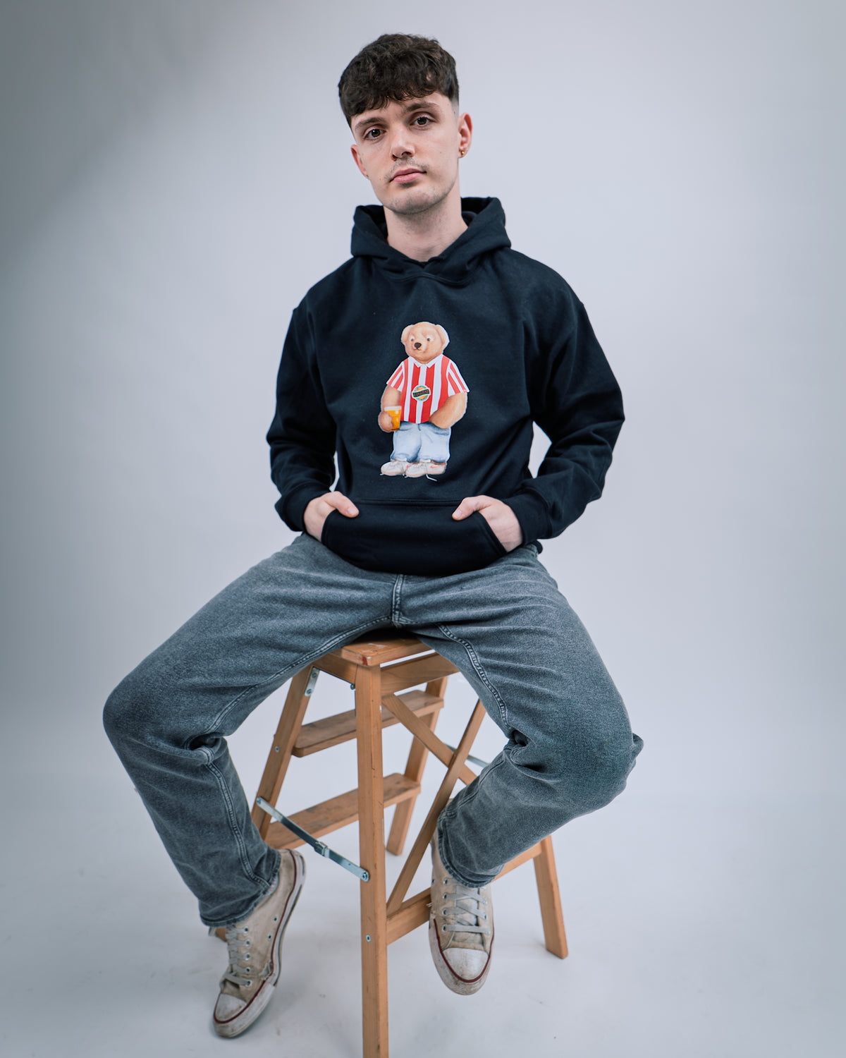 Person wearing a black hoodie with a teddy bear graphic, sitting on a wooden stool against a plain background.