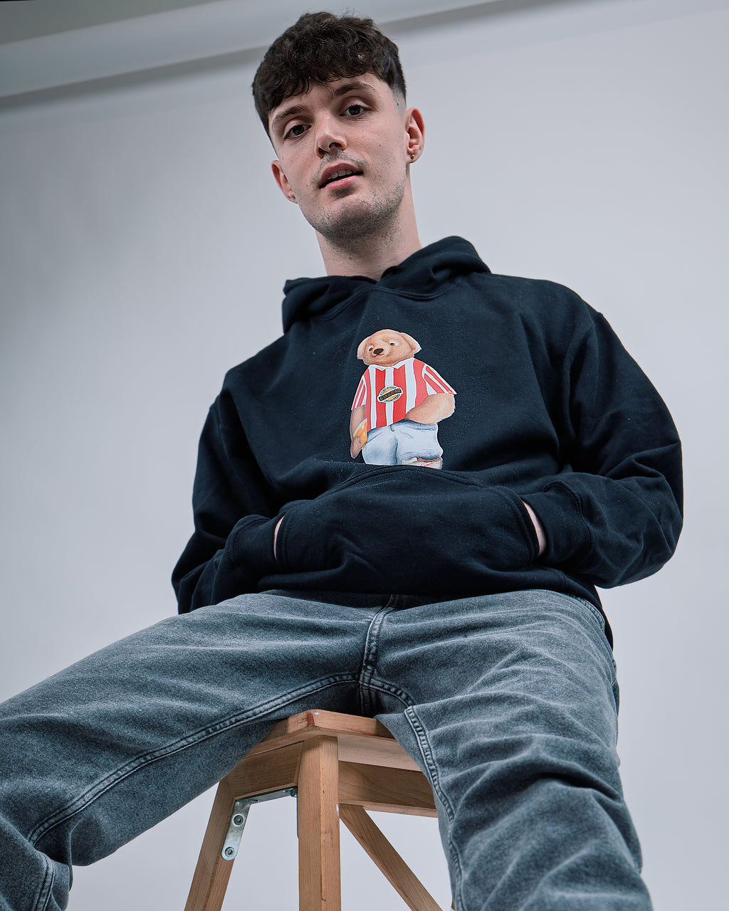 Person wearing a navy hoodie with a graphic design sitting on a stool against a plain background