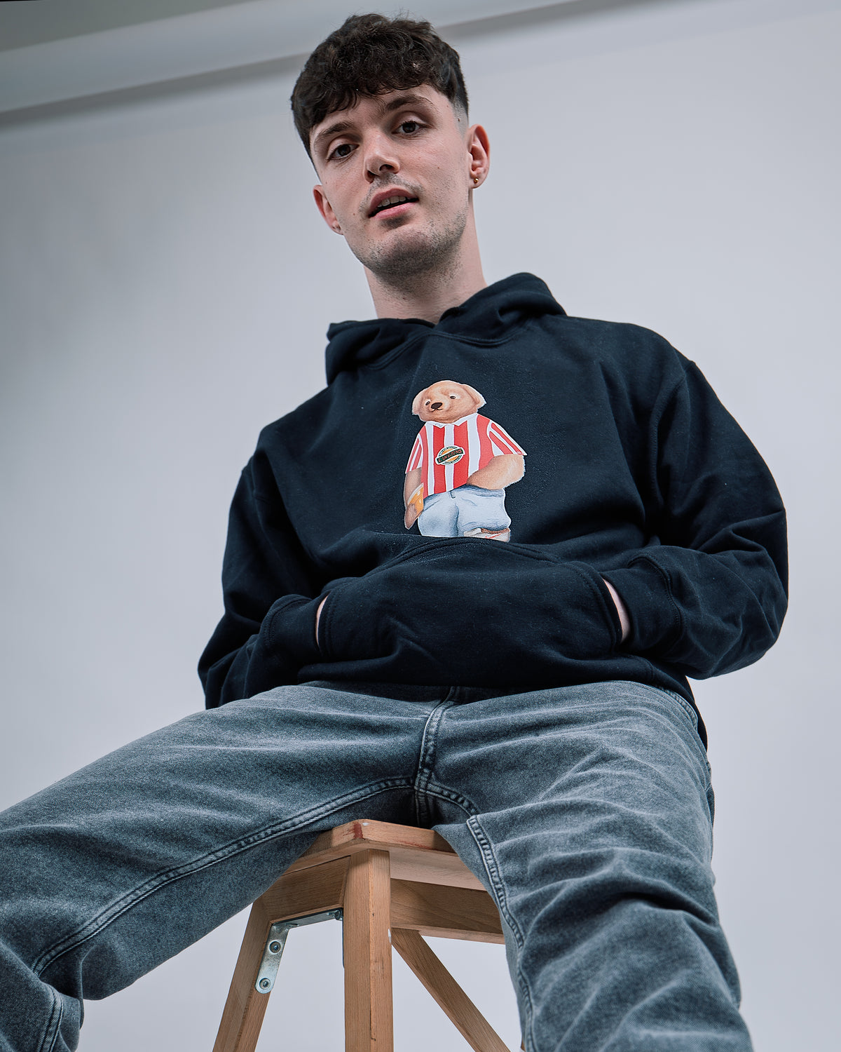 Person wearing a navy hoodie with a graphic design sitting on a stool against a plain background