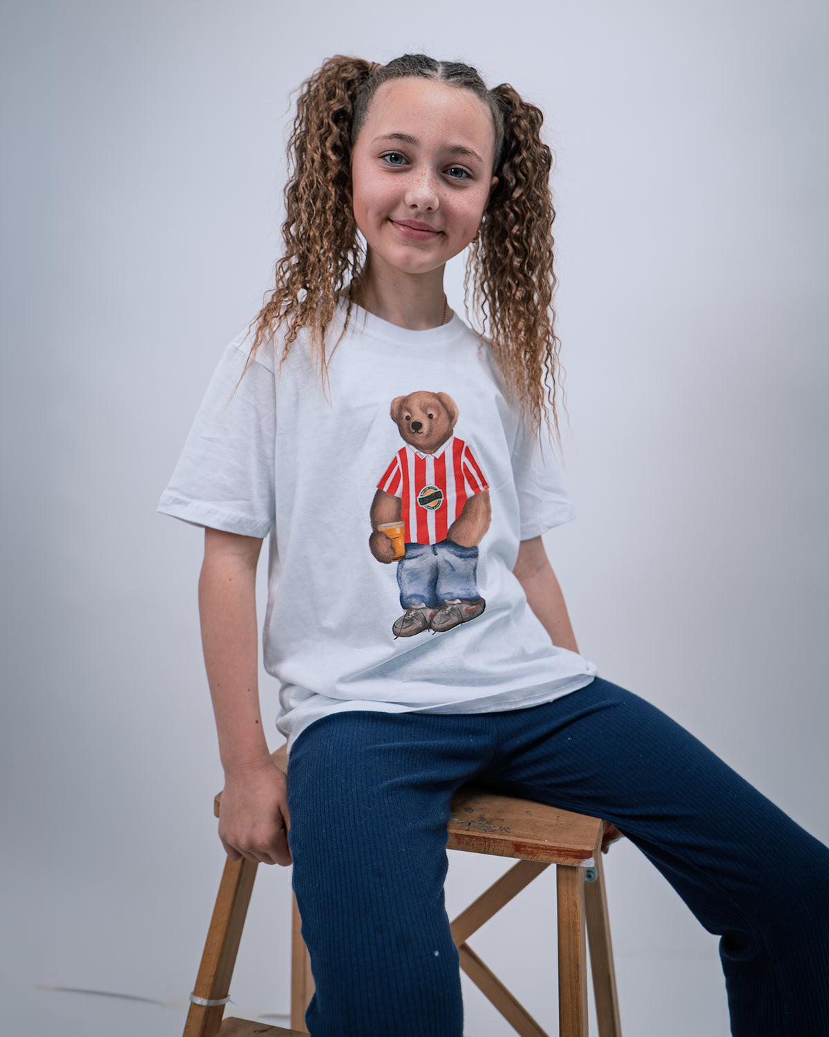 Child wearing a white t-shirt with a bear graphic sitting on a stool against a plain background