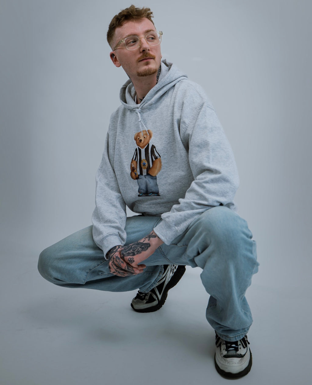 Man wearing a gray hoodie with a bear graphic and light blue jeans on a gray background