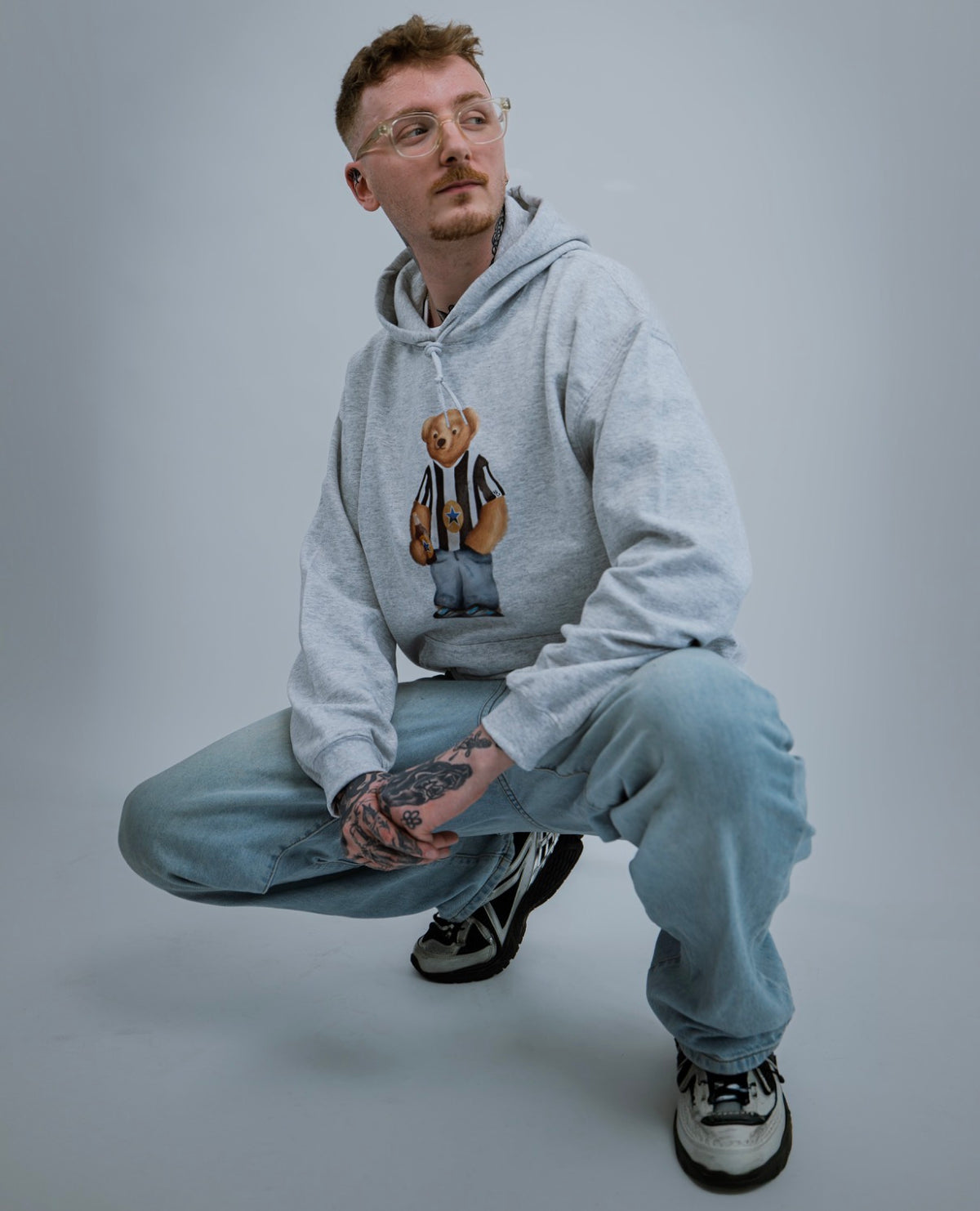 Man wearing a gray hoodie with a bear graphic and light blue jeans on a gray background