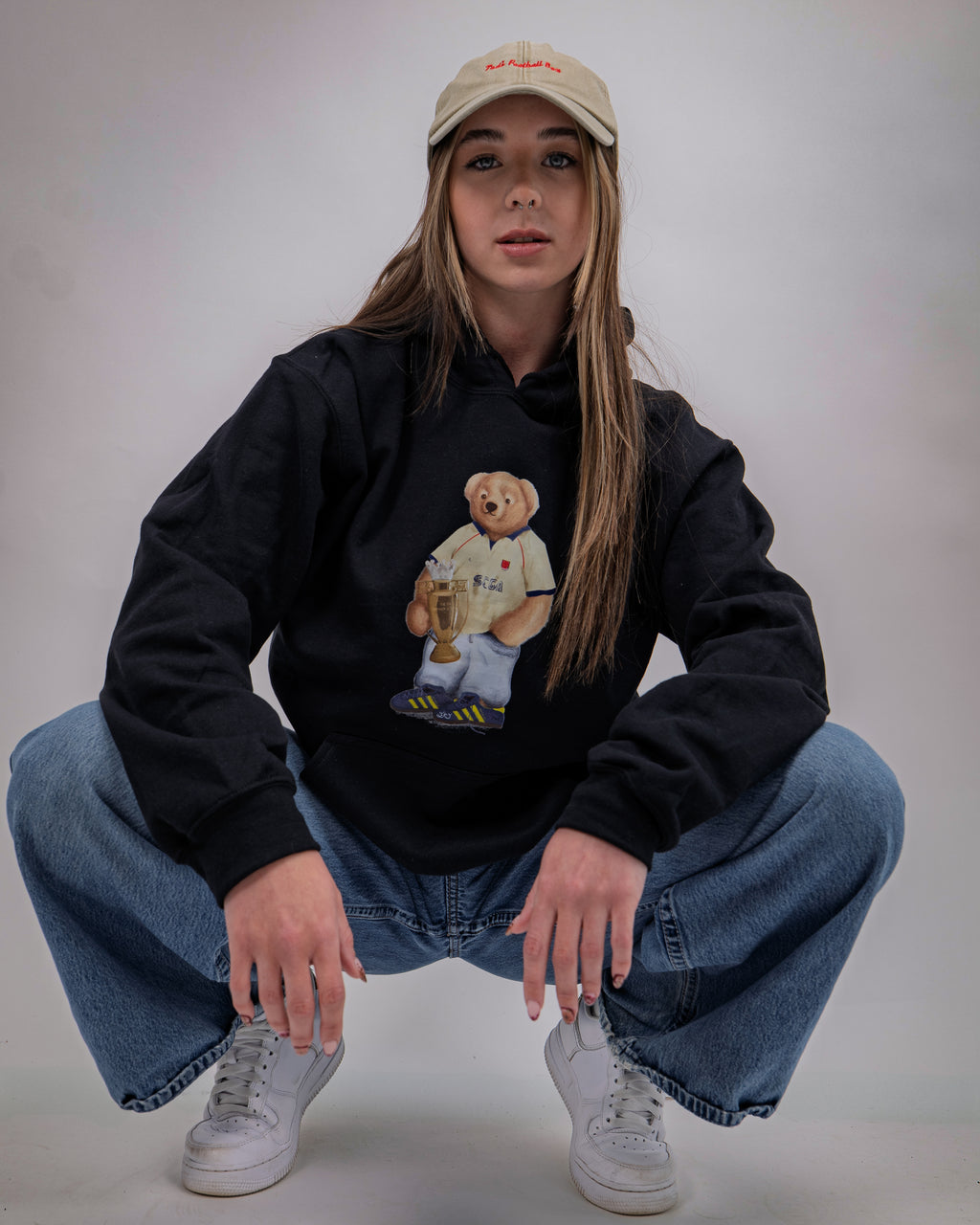 Person wearing a black hoodie with a teddy bear graphic, blue jeans, and a beige cap on a plain background