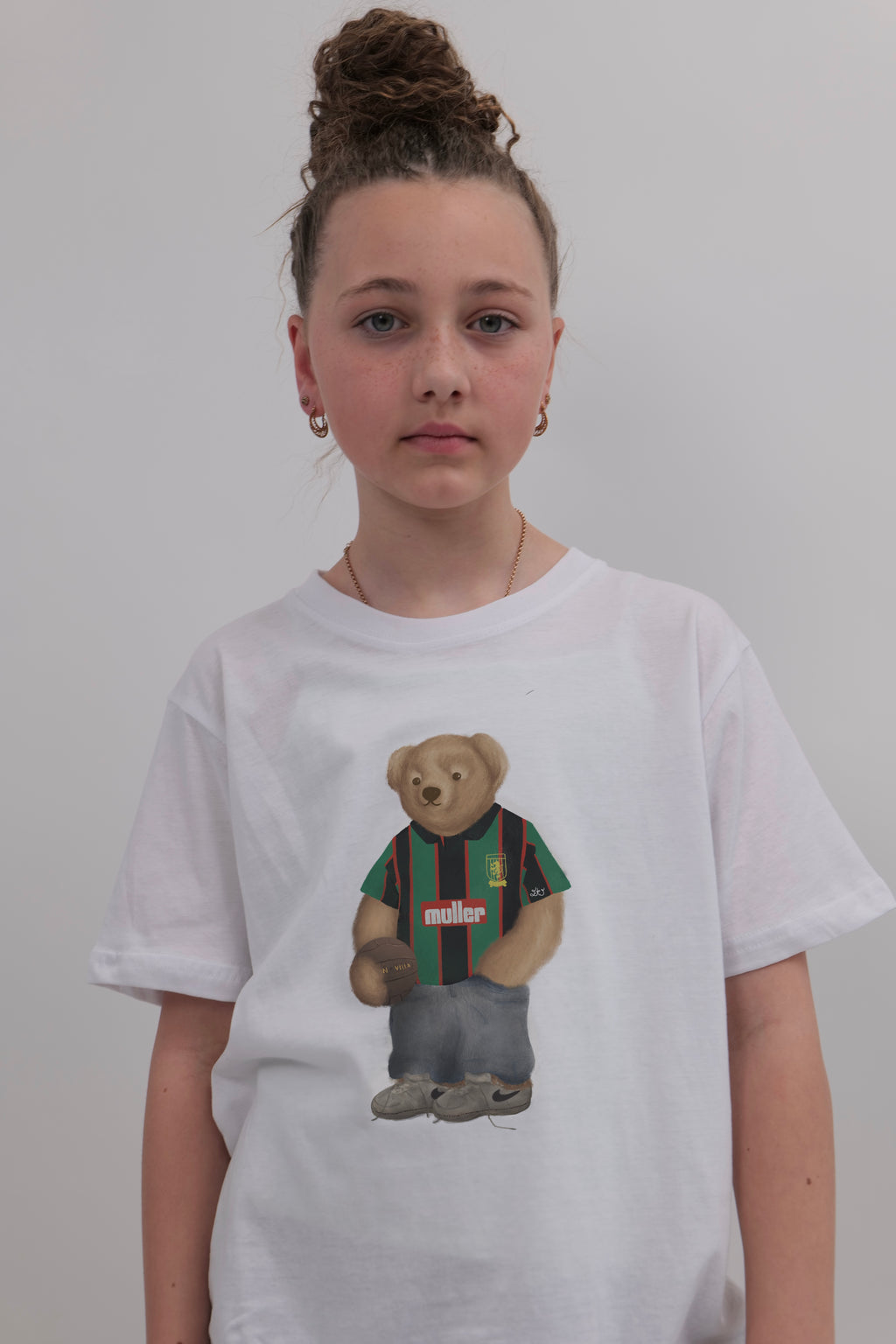 A young girl with her hair in a high bun wears a white crewneck t-shirt featuring a graphic of a teddy bear wearing a green and red striped jersey. The background is a plain light gray wall.