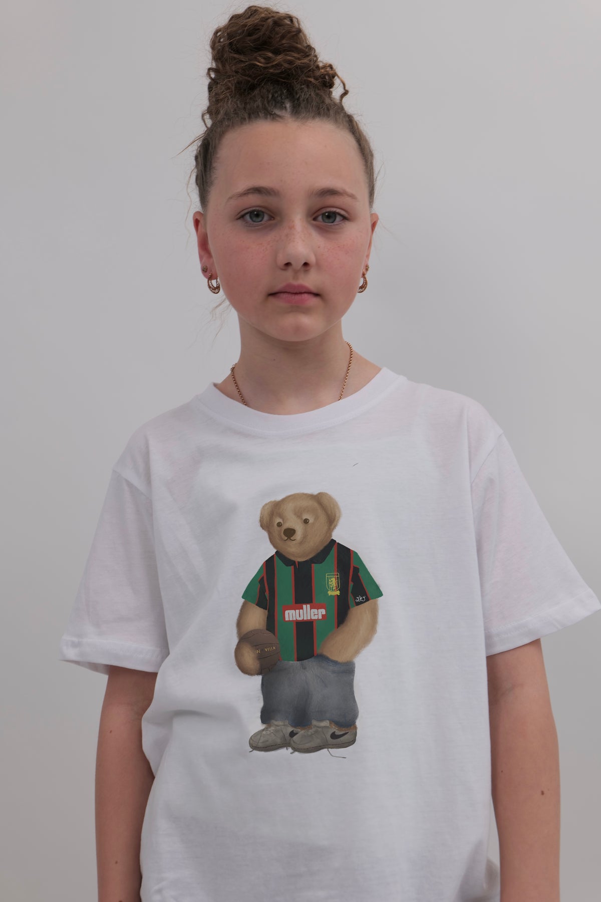 A young girl with her hair in a high bun wears a white crewneck t-shirt featuring a graphic of a teddy bear wearing a green and red striped jersey. The background is a plain light gray wall.