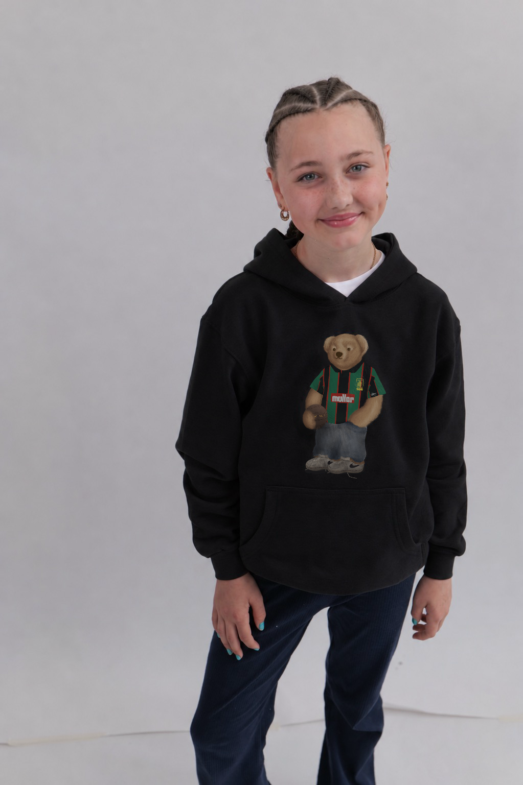 A child wearing a black pullover hoodie with a teddy bear graphic on the front, paired with dark blue pants, standing against a light gray studio background.