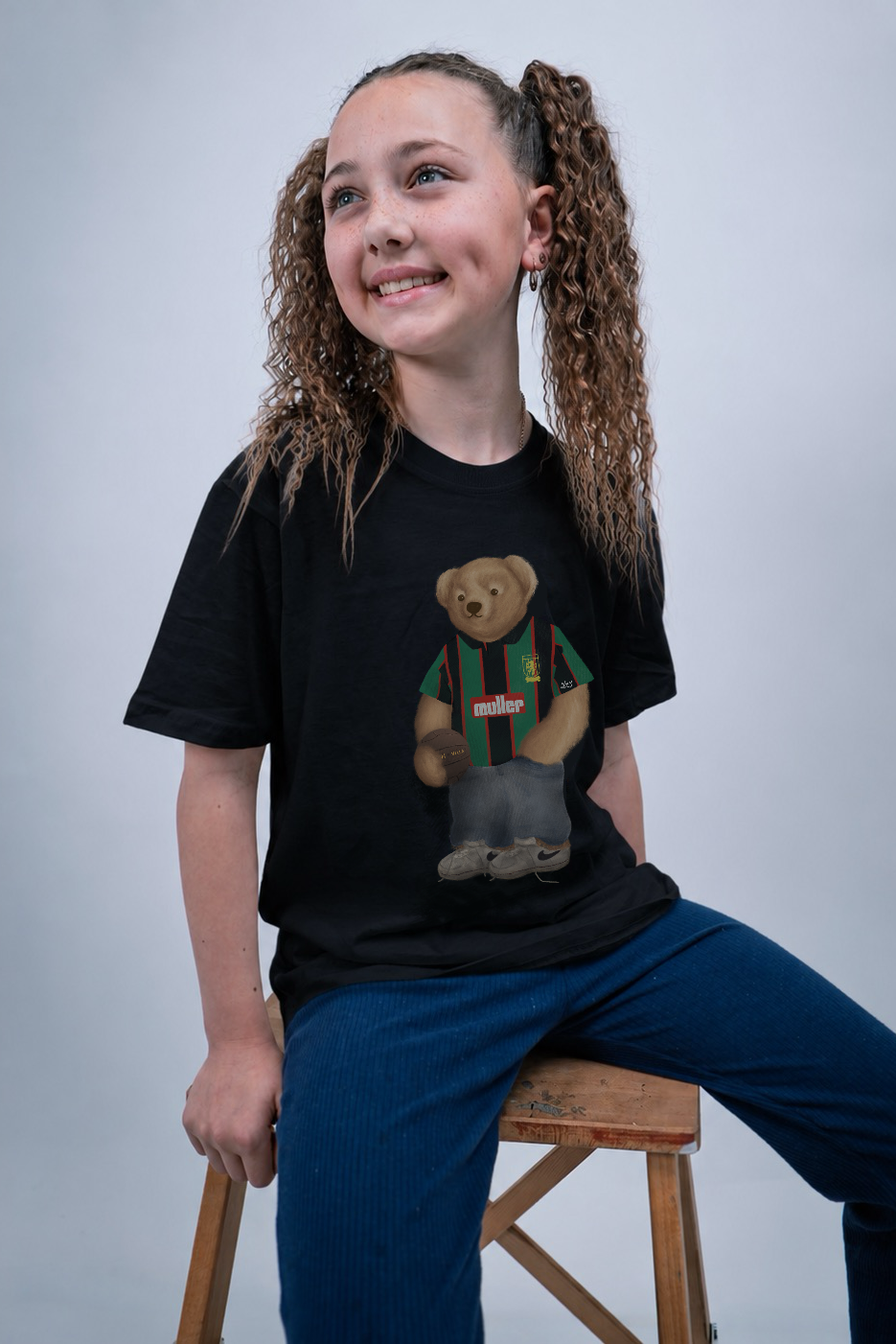 A smiling girl with curly brown hair in pigtails sits on a wooden stool wearing a black graphic t-shirt with a teddy bear wearing green and red striped clothing, paired with blue pants, against a light gray studio background.
