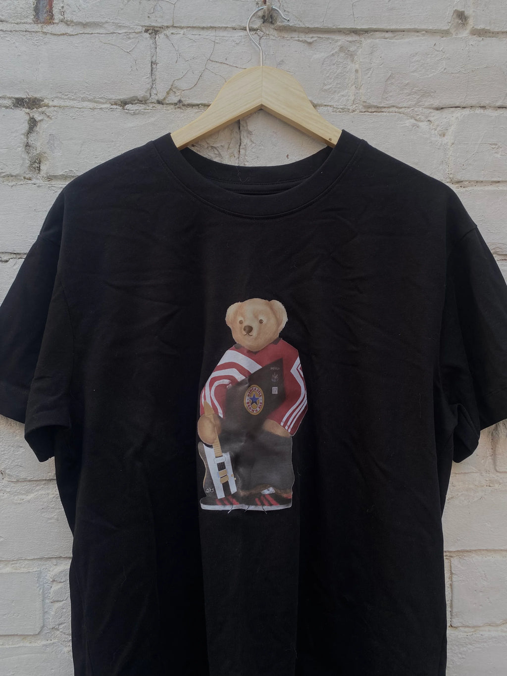 Black t-shirt with a graphic of a bear wearing a striped shirt on a hanger against a white brick wall.