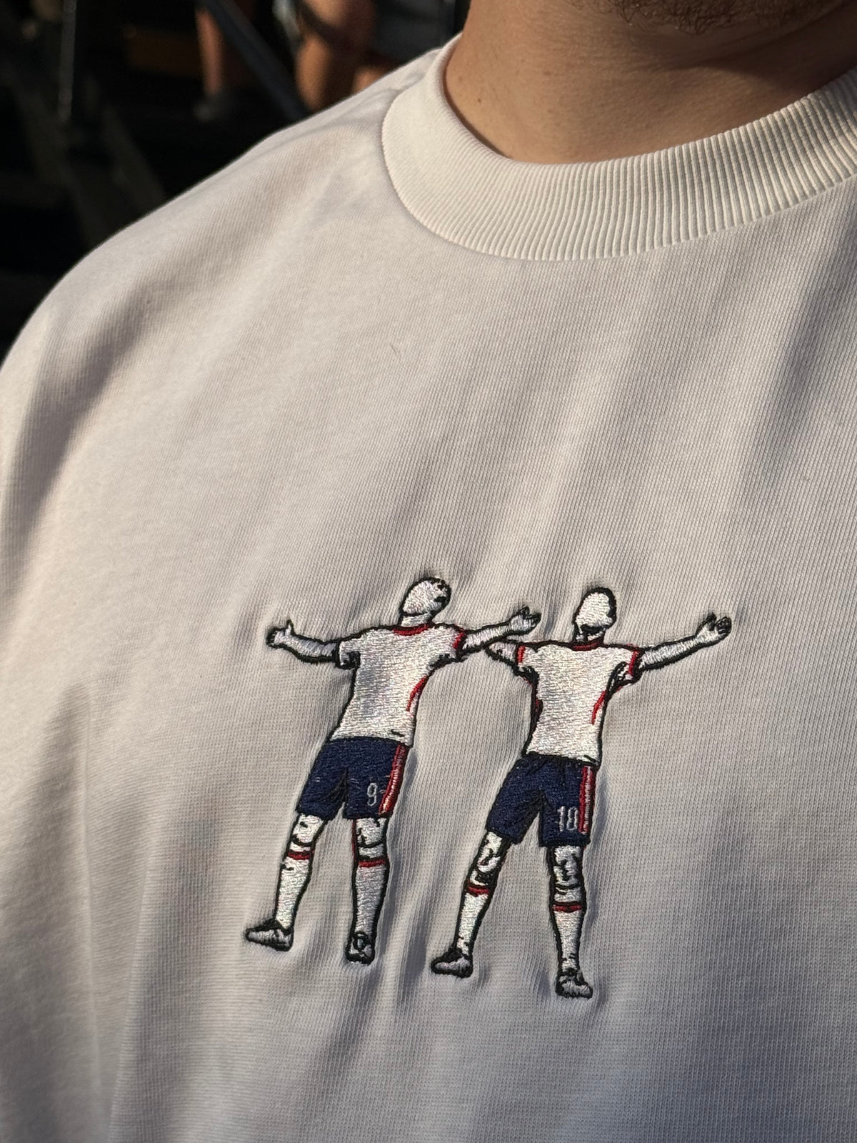Close-up of a white sleeveless top or dress with embroidered soccer player figures on the chest, showing part of a person's neck and shoulder.