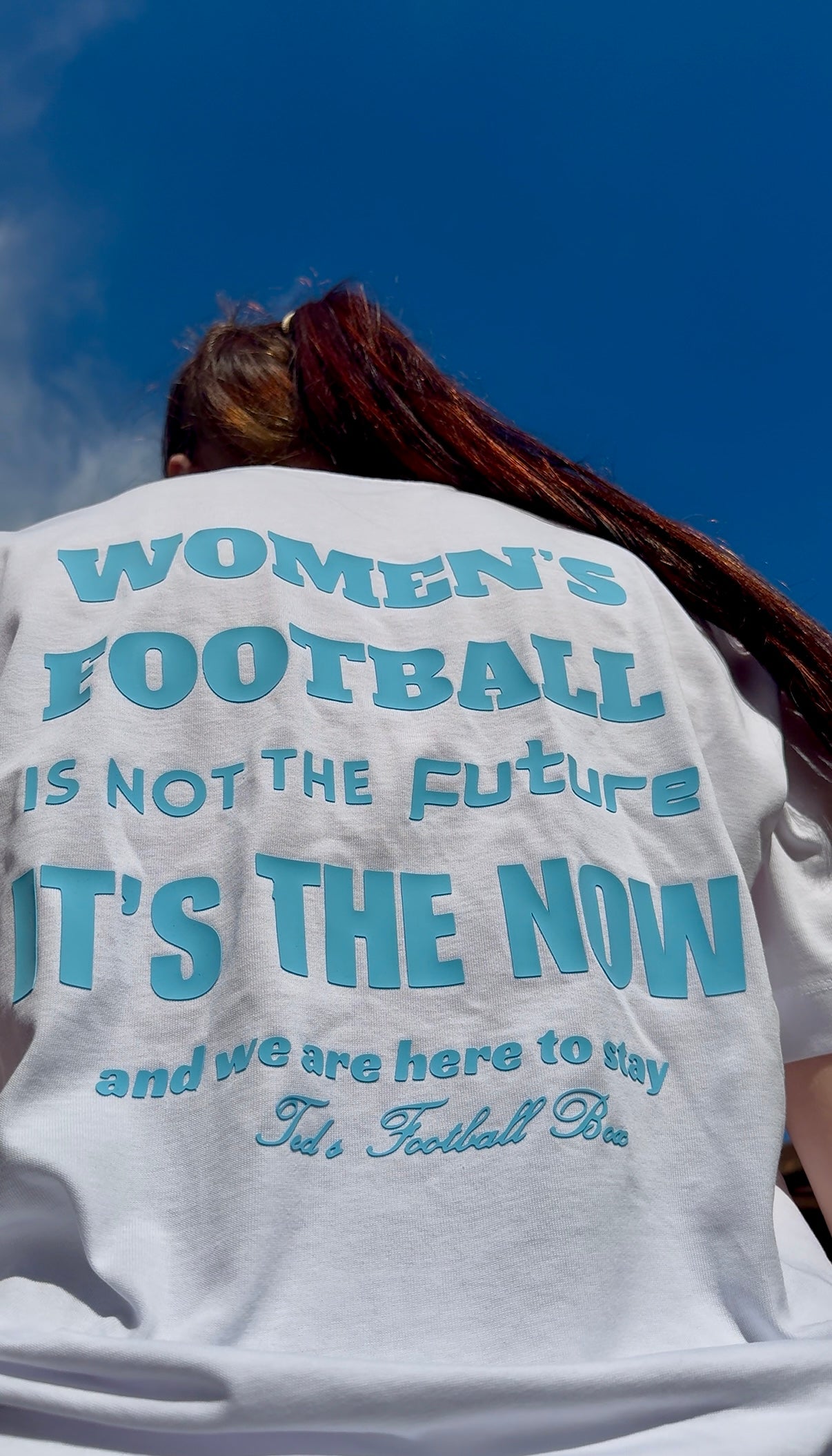 Person wearing a shirt with 'Women's Football is not the future, it's the now' text against a blue sky.