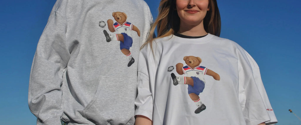 Two people wearing teds tshirts and hoodies with a bear playing soccer design against a clear blue sky