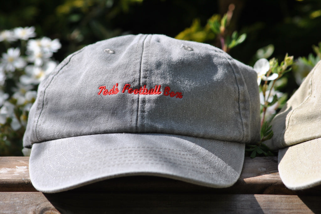 Gray cap with red  embroidered text 'teds football box'on a wooden surface with greenery in the background