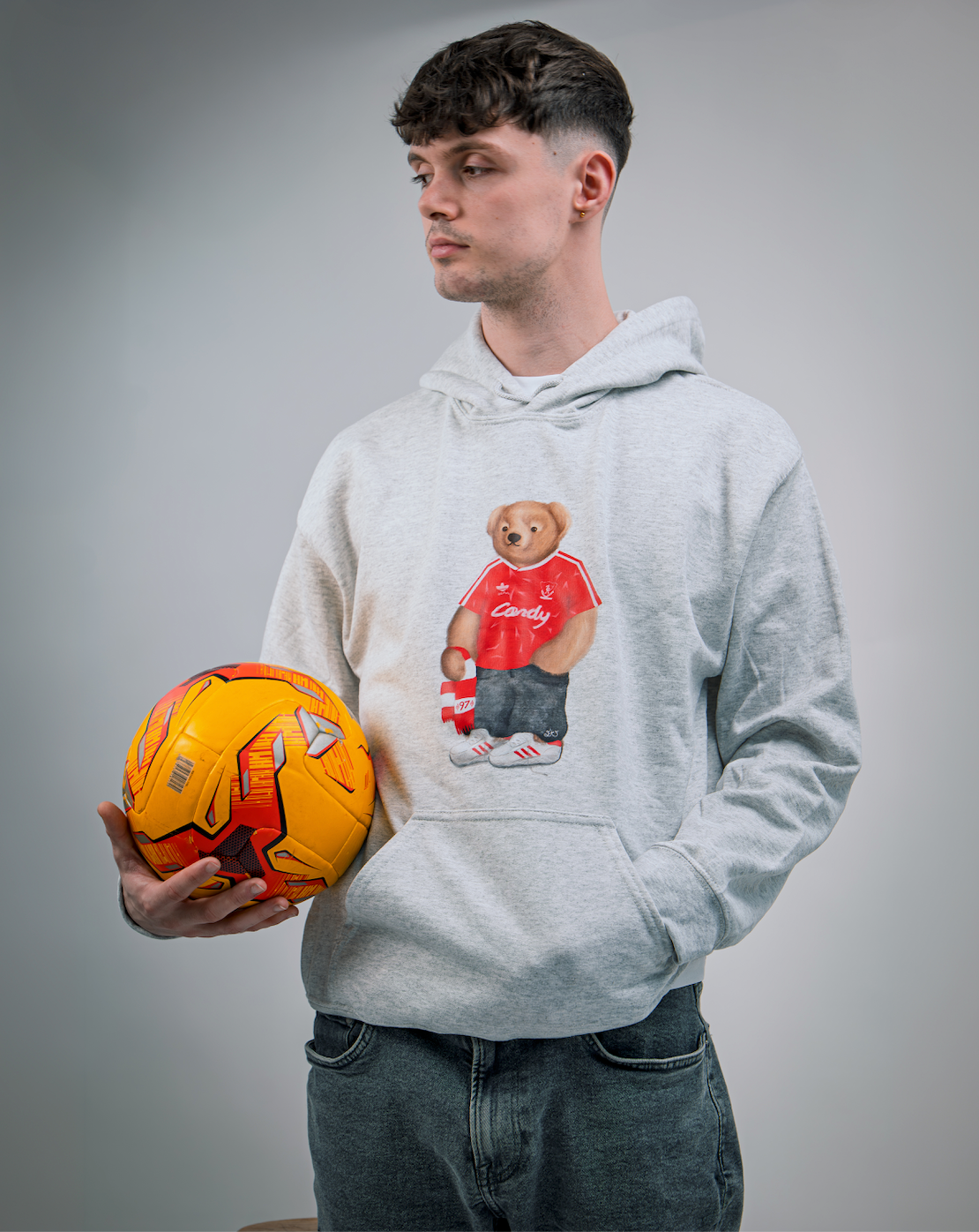 Person wearing a hoodie with a bear graphic holding an orange soccer ball on a gray background