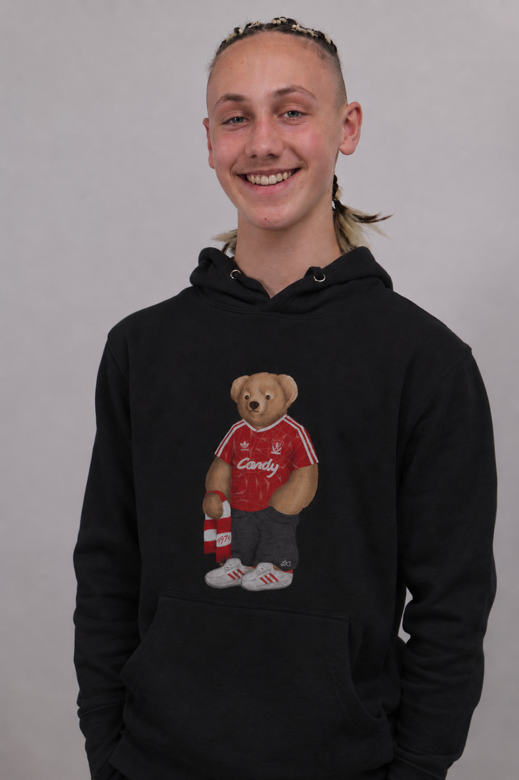 A smiling young person wearing a black pullover hoodie with a teddy bear graphic on the chest. The graphic shows a brown teddy bear in a red jersey holding a red and white striped drink can.