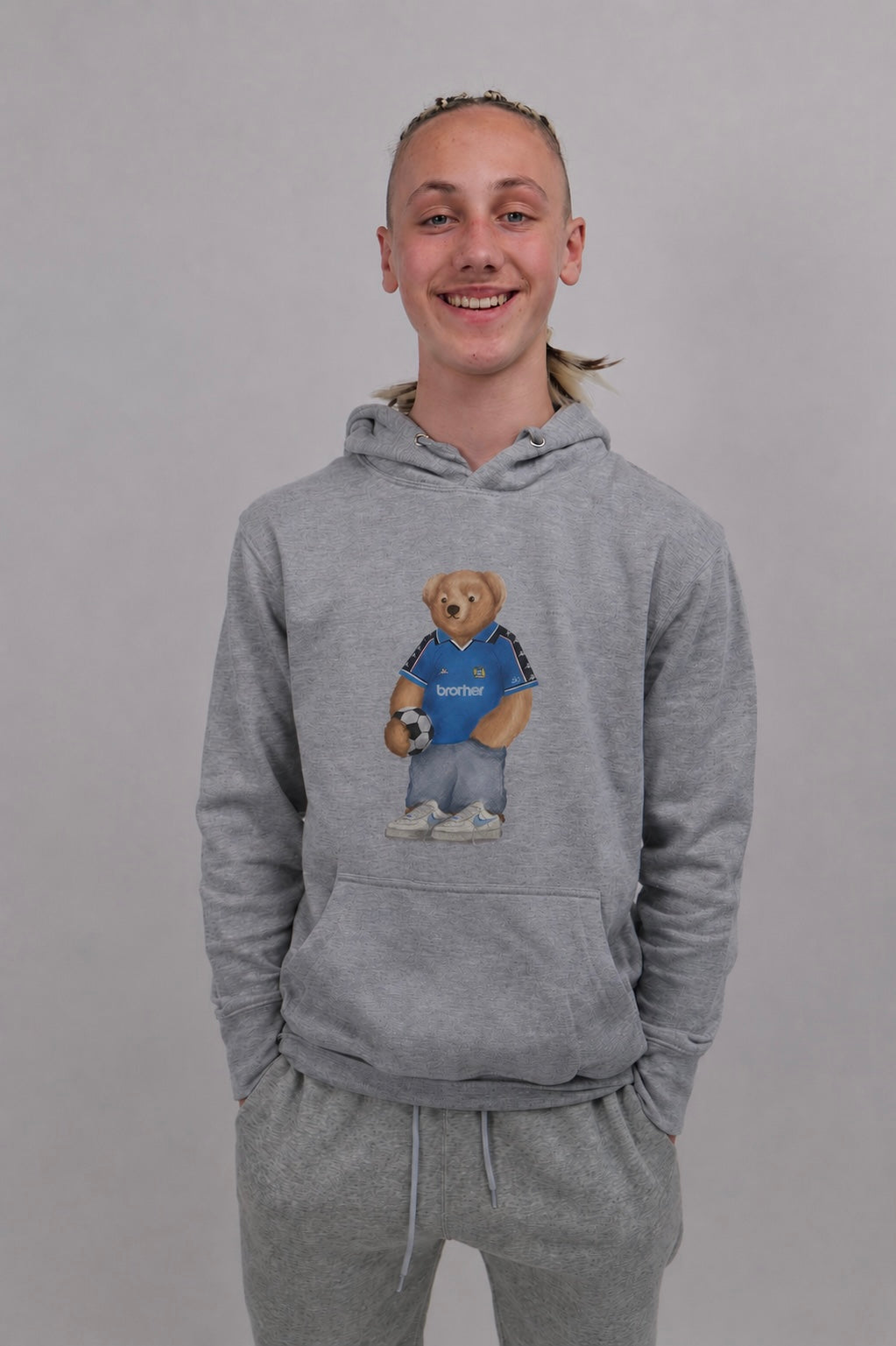 A smiling young man wearing a heather gray pullover hoodie with a teddy bear graphic holding a soccer ball, paired with matching gray sweatpants, against a plain gray studio background.