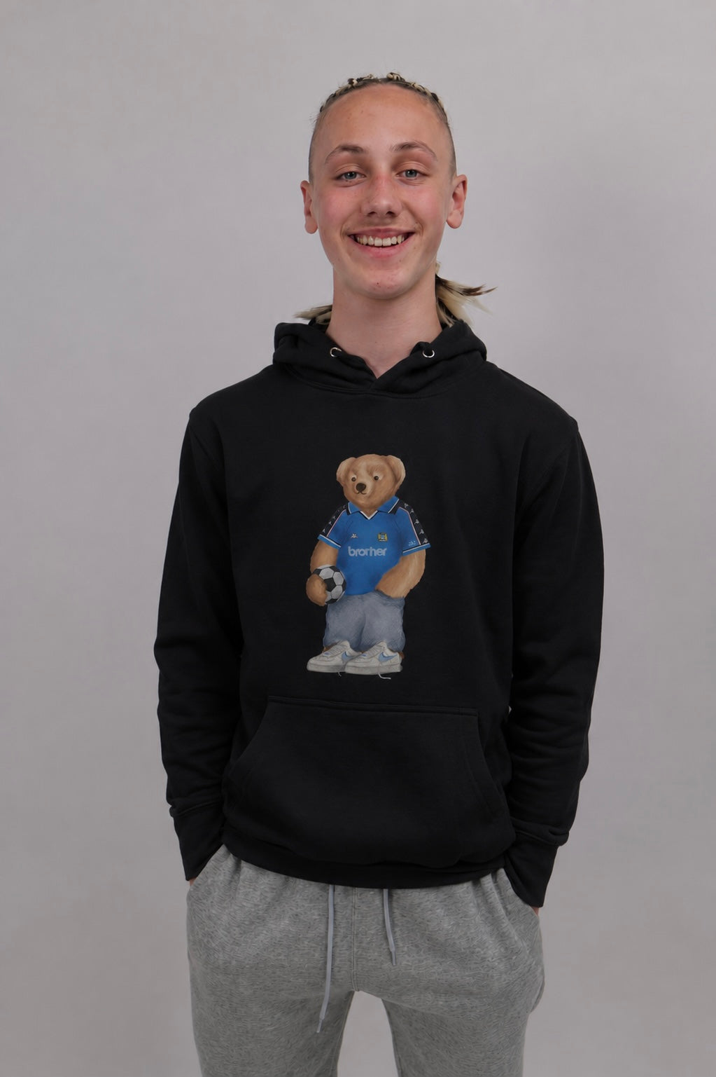 A smiling young man wearing a black pullover hoodie with a teddy bear graphic on the chest, paired with gray sweatpants, standing against a plain light gray background.