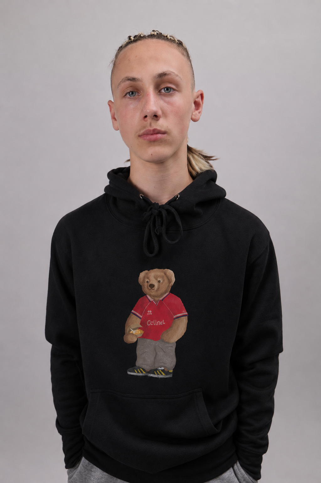 A person wearing a black pullover hoodie with a graphic of a teddy bear in a red shirt holding a cup, standing against a plain gray background.