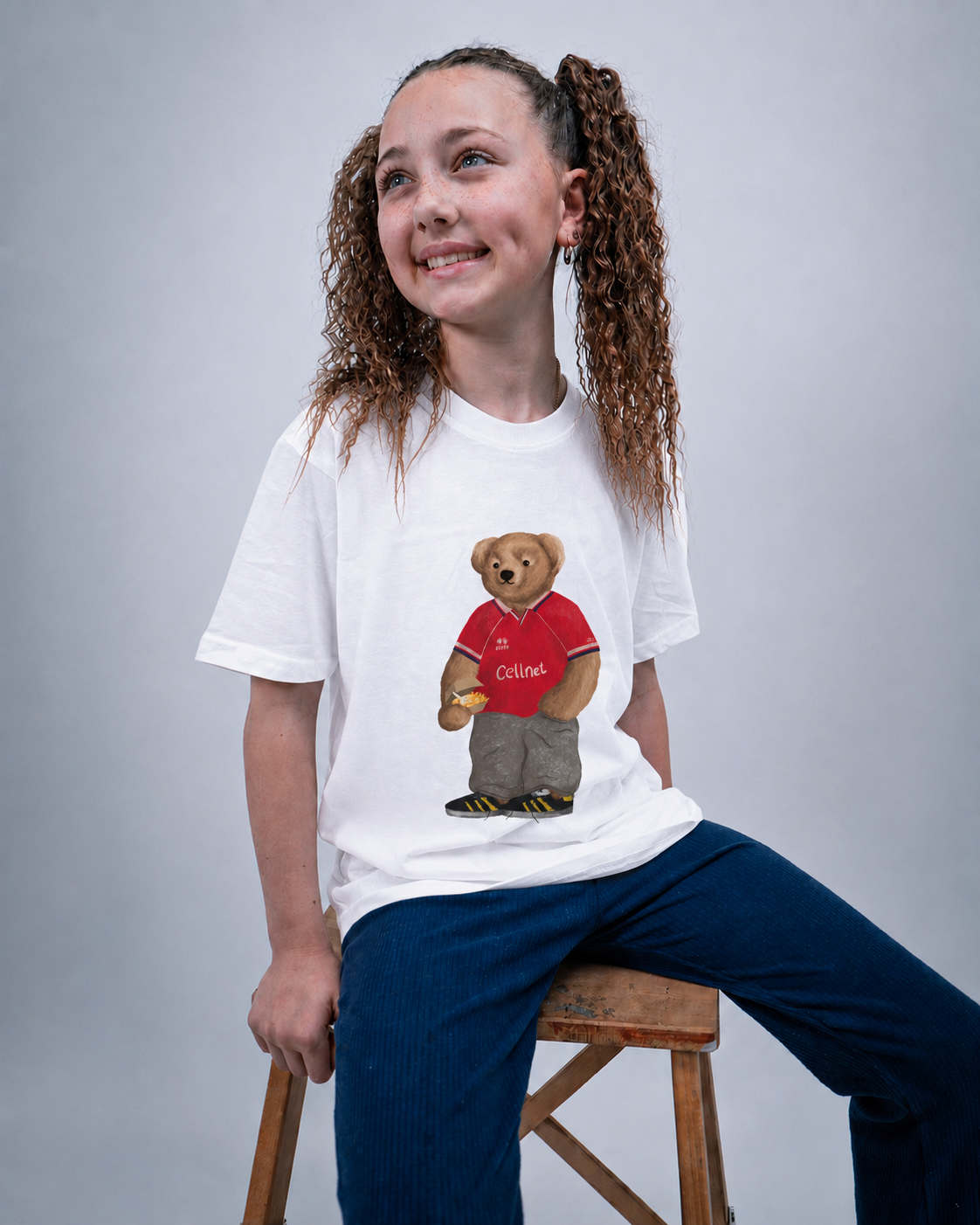 A smiling girl with curly brown hair in pigtails sits on a wooden stool, wearing a white t-shirt with a graphic of a teddy bear in a red Collins jersey and navy blue pants against a gray studio background.