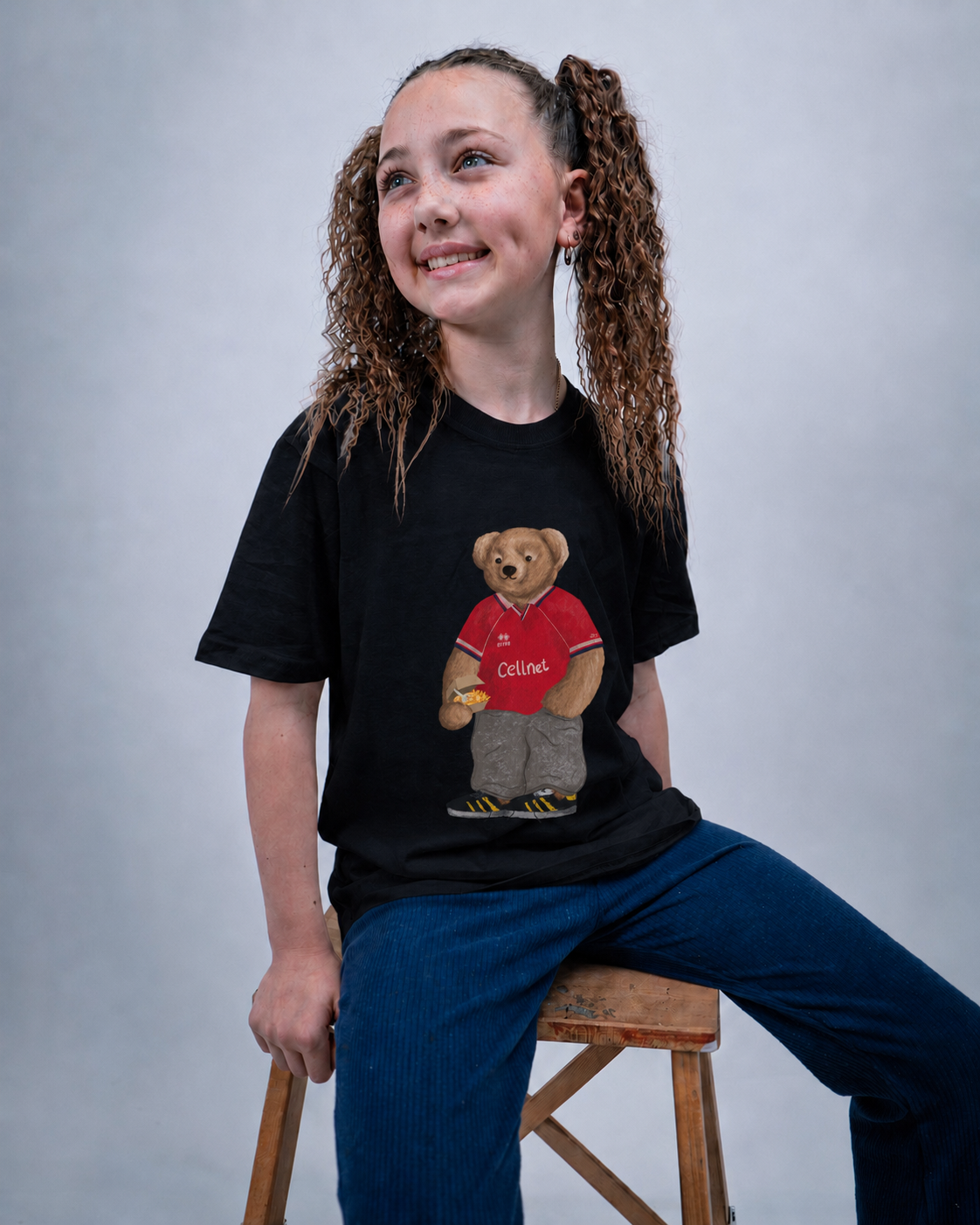 A smiling girl with curly brown hair in pigtails sits on a wooden stool, wearing a black t-shirt with a graphic of a teddy bear in a red Collins jersey and blue jeans against a gray studio background.