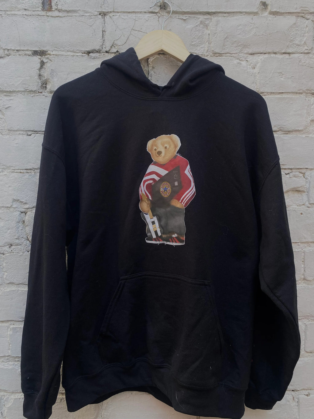 Black hoodie with a graphic a bear on a hanger against a brick wall.
