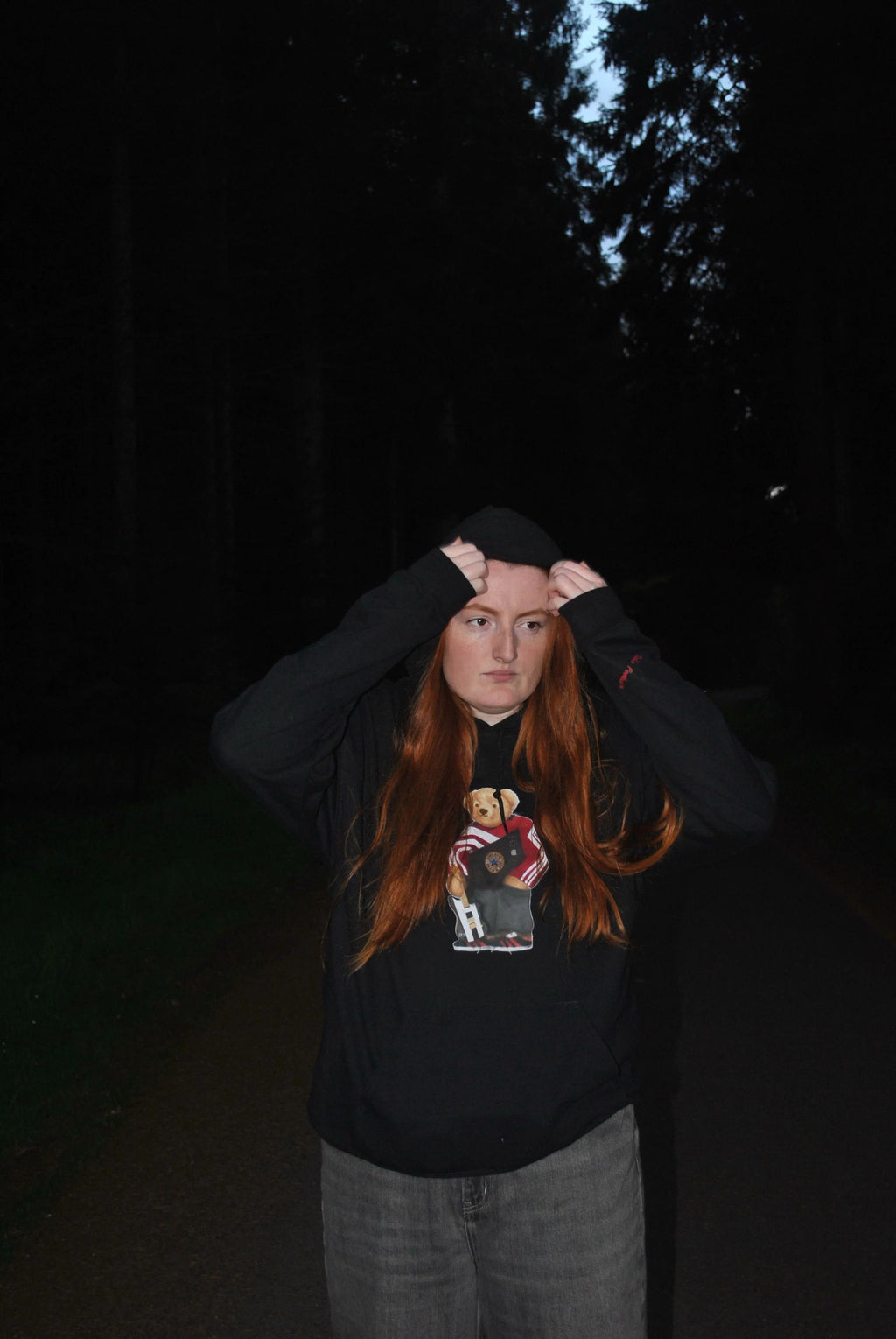 Person with long red hair wearing a black hoodie with a graphic design, standing in a dark forest.