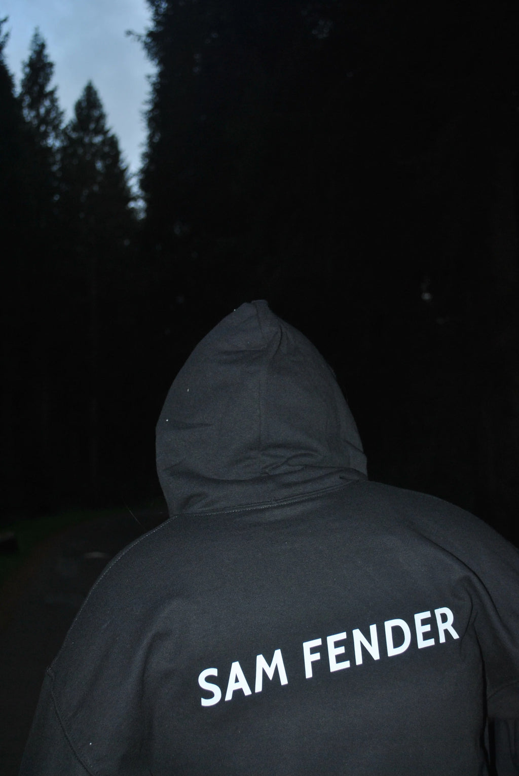 Person wearing a hoodie with 'SAM FENDER' text in a dark forest setting