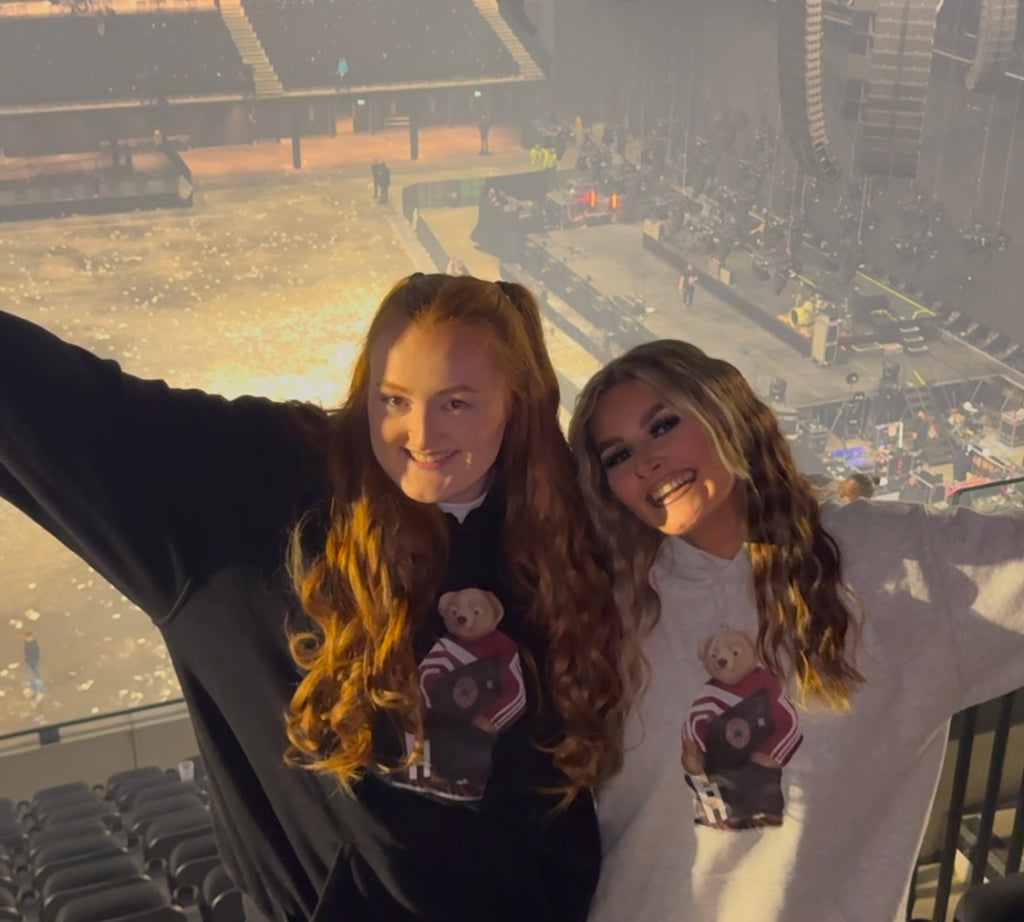 Two women wearing matching hoodies with teddy bear designs in a stadium setting.