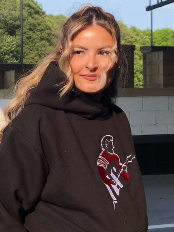 young woman wearing a black hoodie with an embroidered sam fender design 