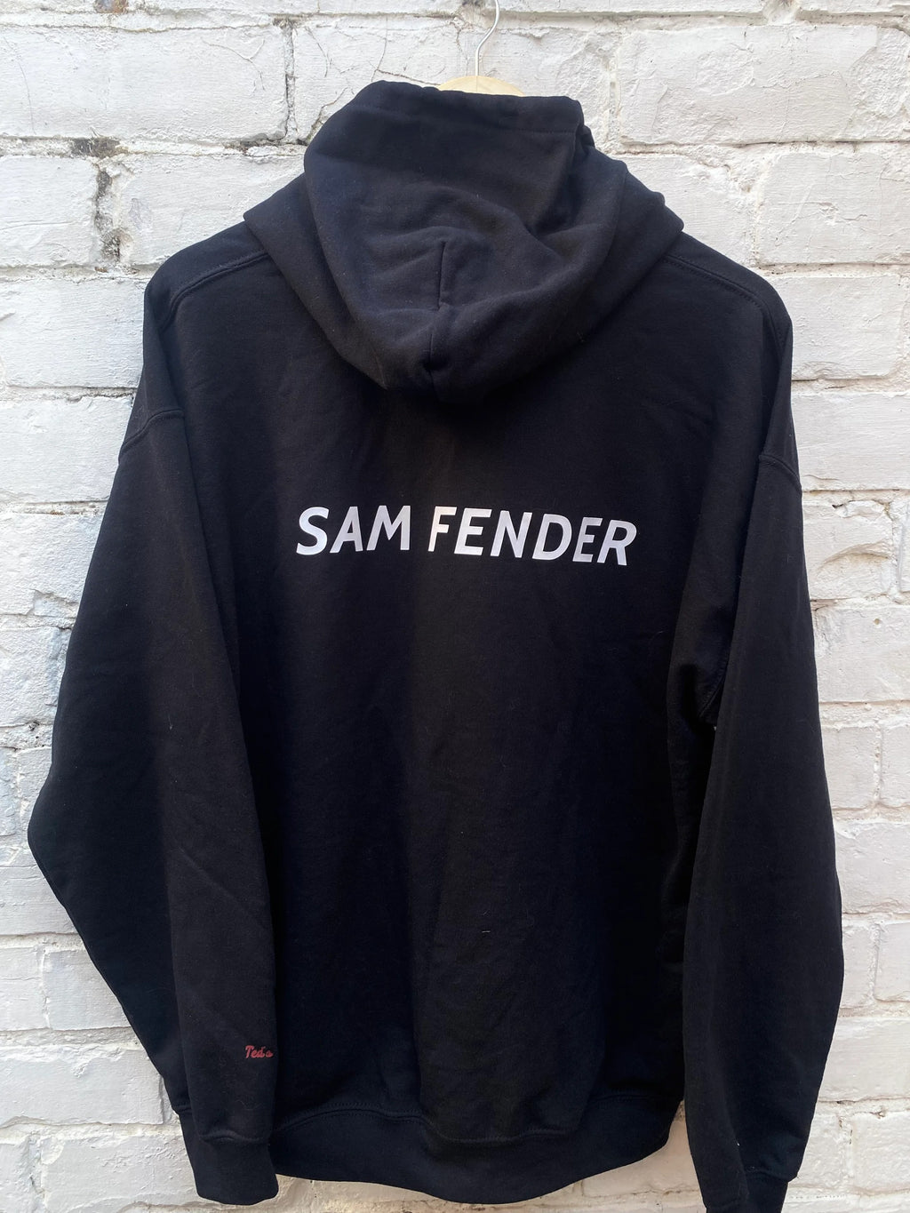 Black hoodie with 'SAM FENDER' text on the back against a light brick wall.