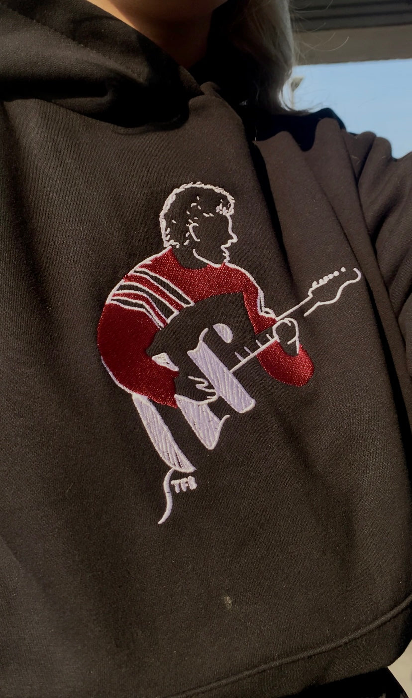 Dark hoodie with embroidered design of a person playing guitar on a light background