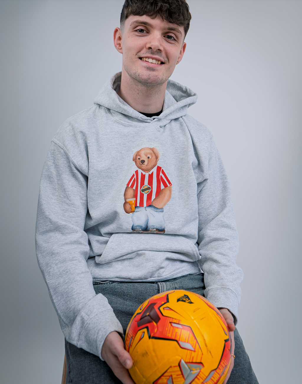 Person wearing a hoodie with a bear graphic holding an orange ball against a gray background