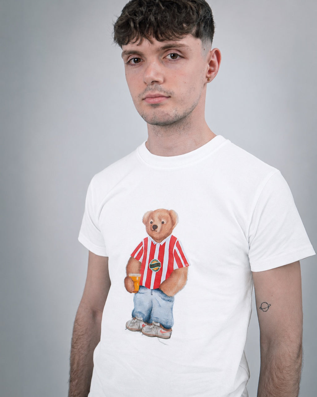 Person wearing a white t-shirt with a bear graphic holding a drink, standing against a gray background