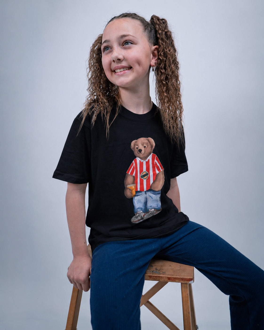 Girl wearing a black t-shirt with a teddy bear graphic, sitting on a wooden stool against a plain background.