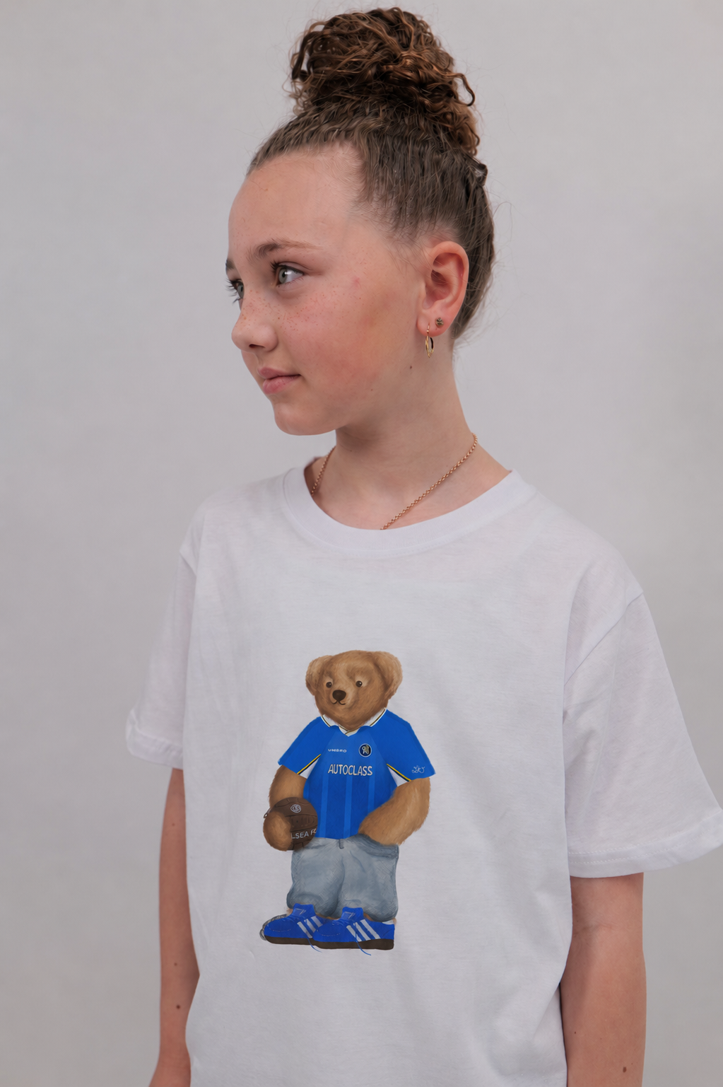 A child wearing a white short-sleeve t-shirt with a graphic of a teddy bear in a blue jersey holding a football, set against a plain light gray background.
