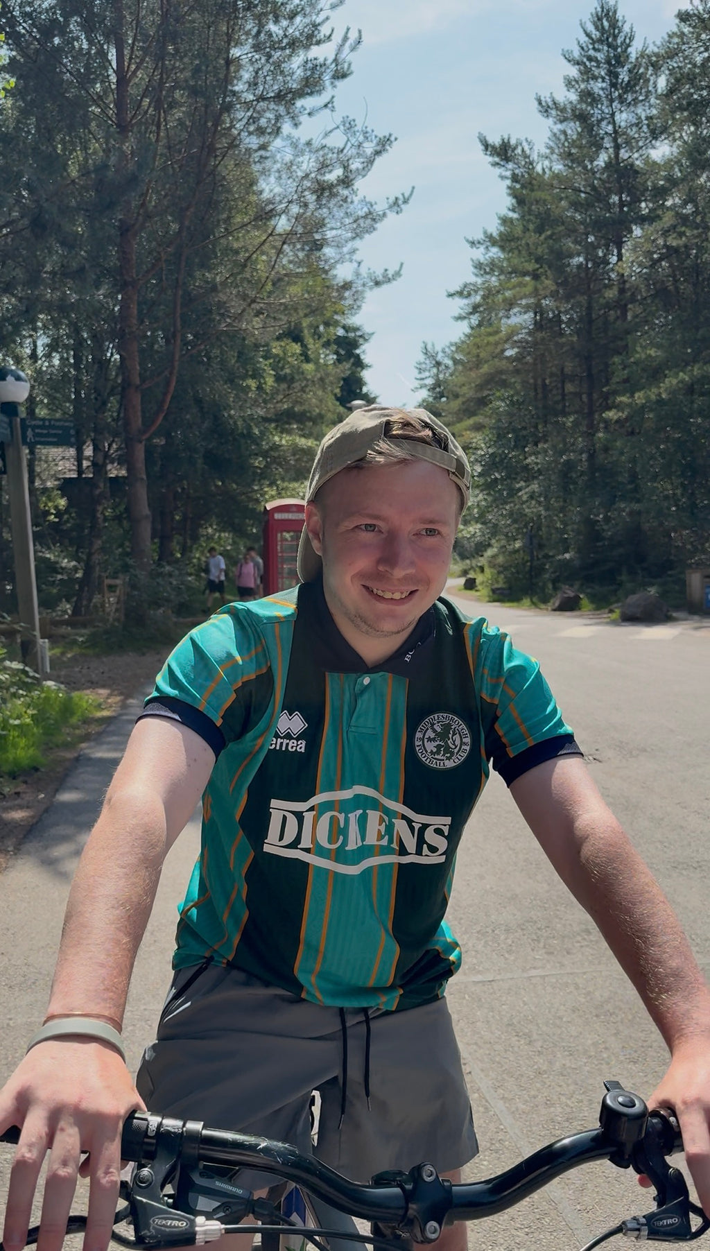 Man wearing a beige cap embroidered with ted 'teds football box" with riding a bicycle on a road with trees in the background, summer time, streetwear