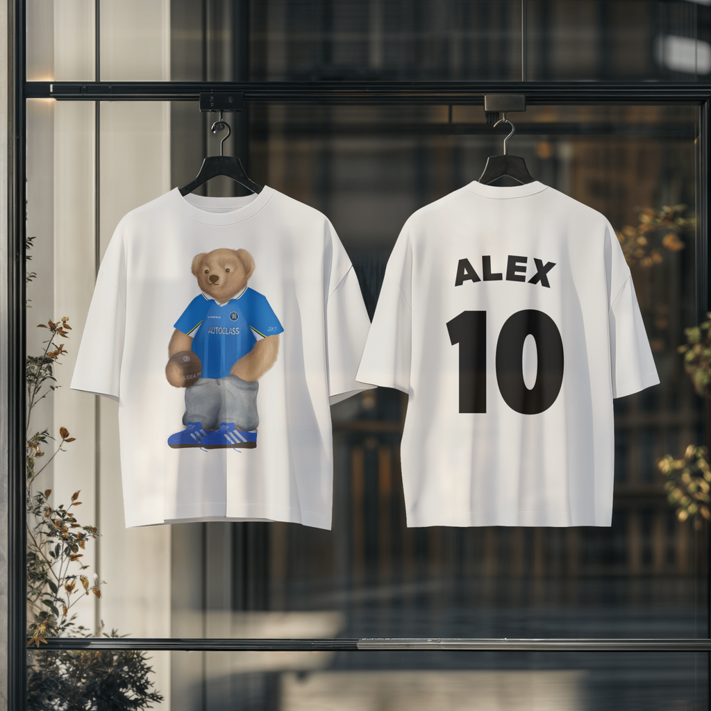 Two white t-shirts with a bear in a blue shirt and 'ALEX 10' printed on them, hanging against a blurred indoor background.